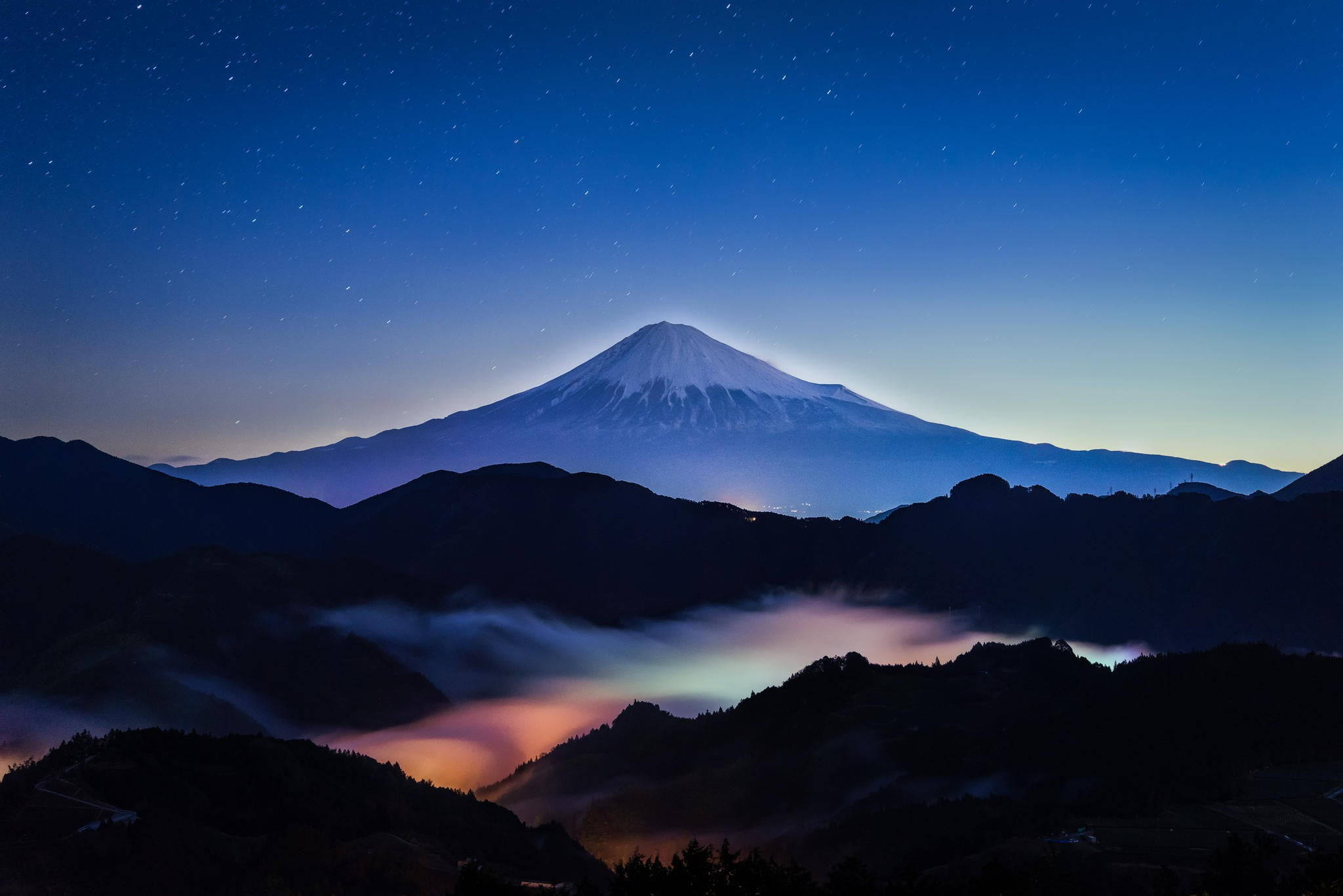 富士山 桌布 壁紙 — 白雪覆蓋的富士山在繁星點點的暮色天空下雄偉地升起，下方有發光的雲彩。