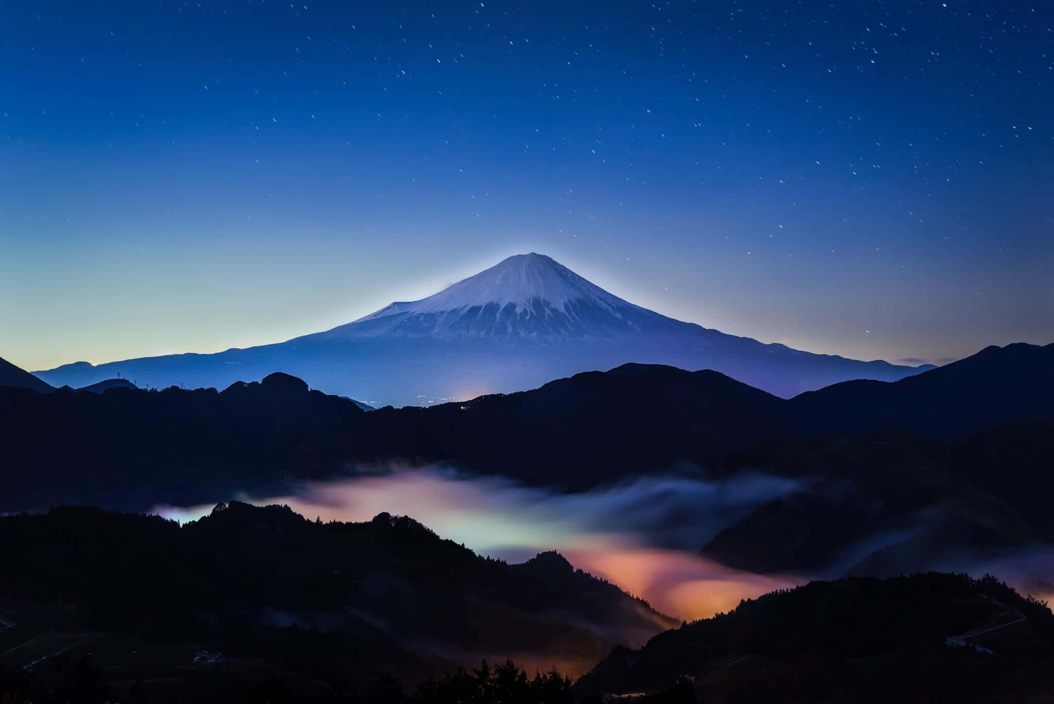 Mt Fuji 桌布 壁紙 — 白雪覆蓋的富士山在繁星點點的暮色天空下巍然聳立，下方有發光的雲彩。