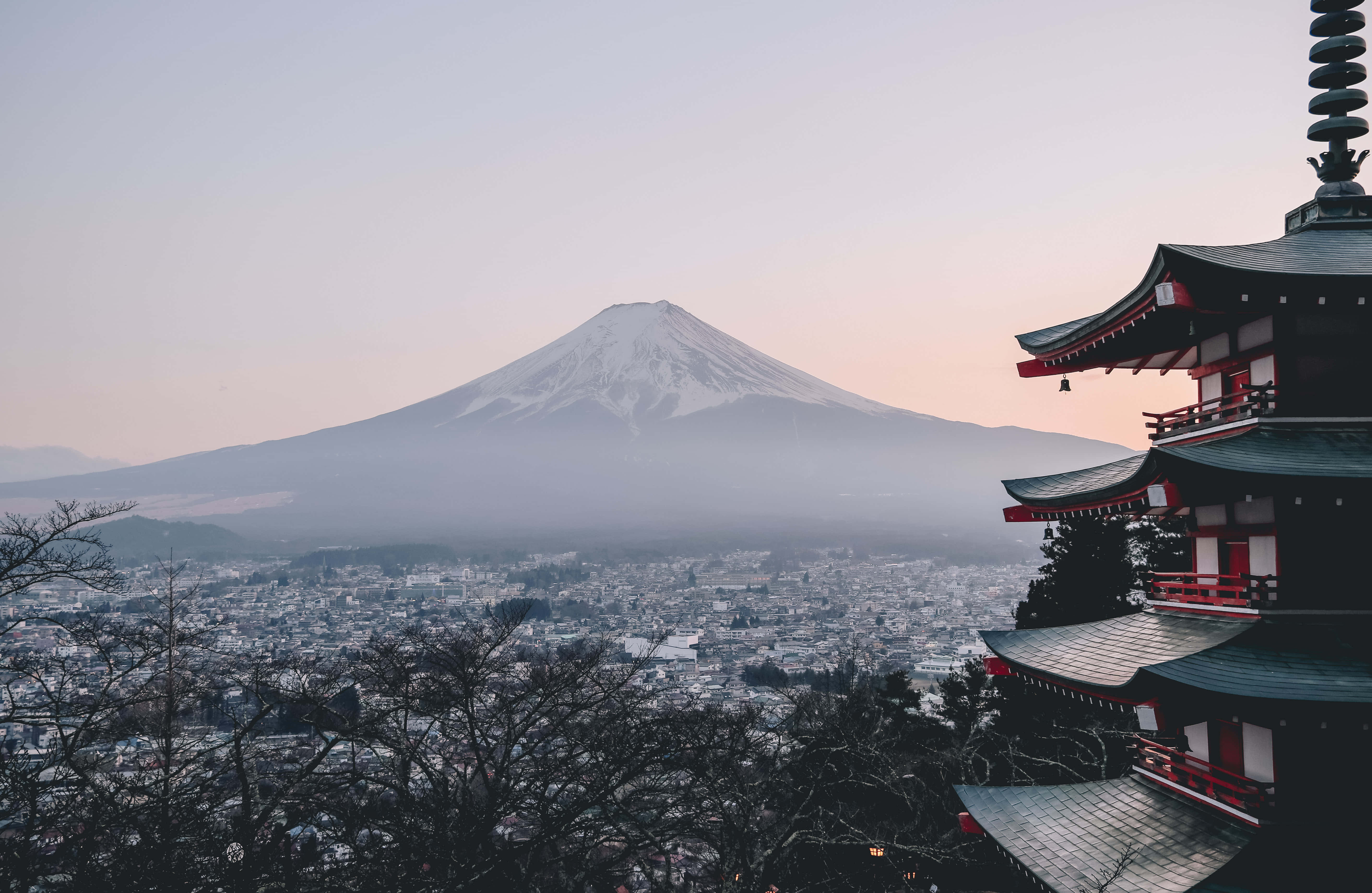 World Travel 桌布 壁紙 — 白雪覆蓋的富士山在霧氣瀰漫的城市景觀後方雄偉地升起，黎明之光沐浴著一座傳統的紅黑相間的寶塔。