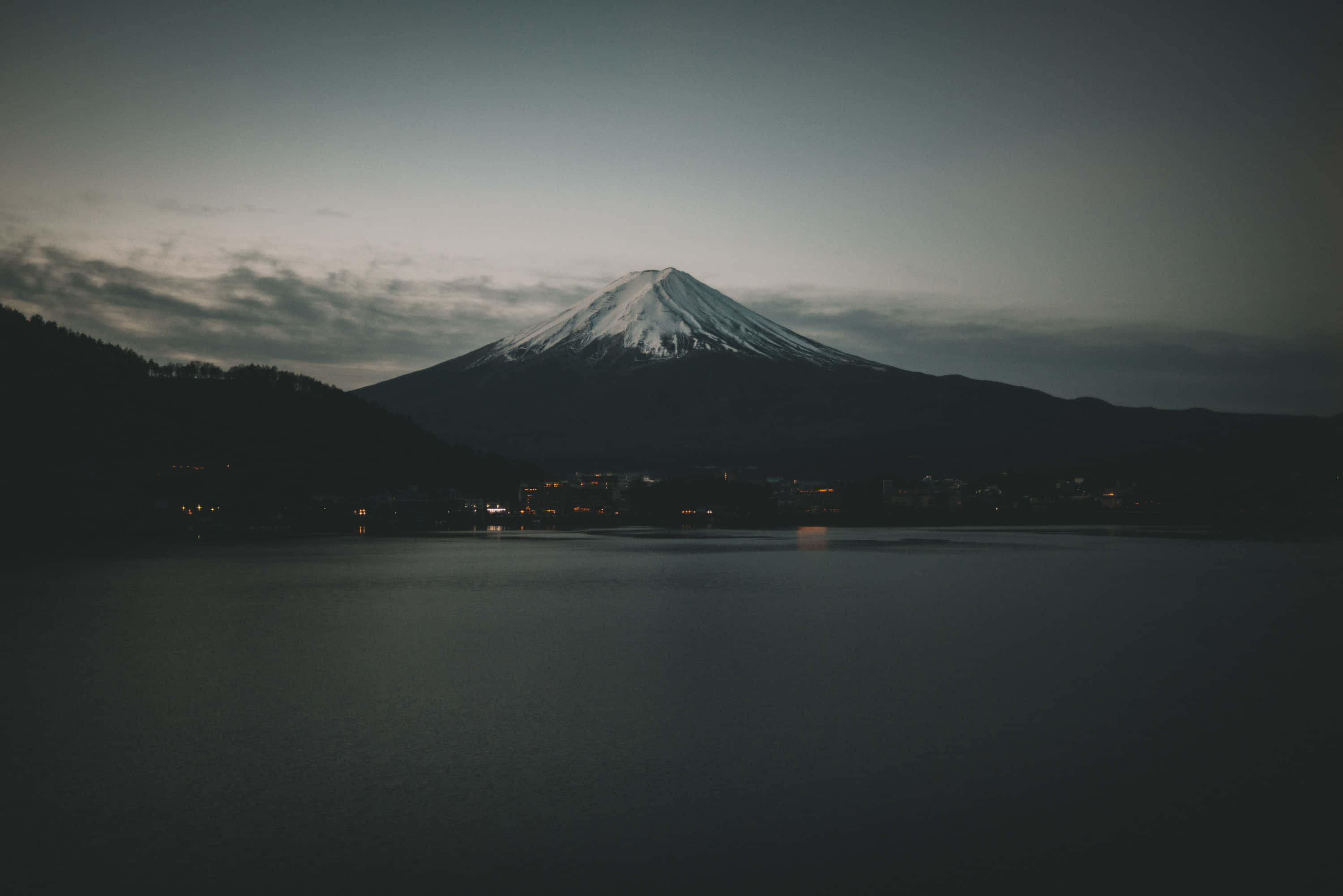 桌布 壁紙 — 白雪覆蓋的富士山在黃昏時分雄偉地聳立在平靜的湖面上，散落的城鎮燈光倒映在水中。