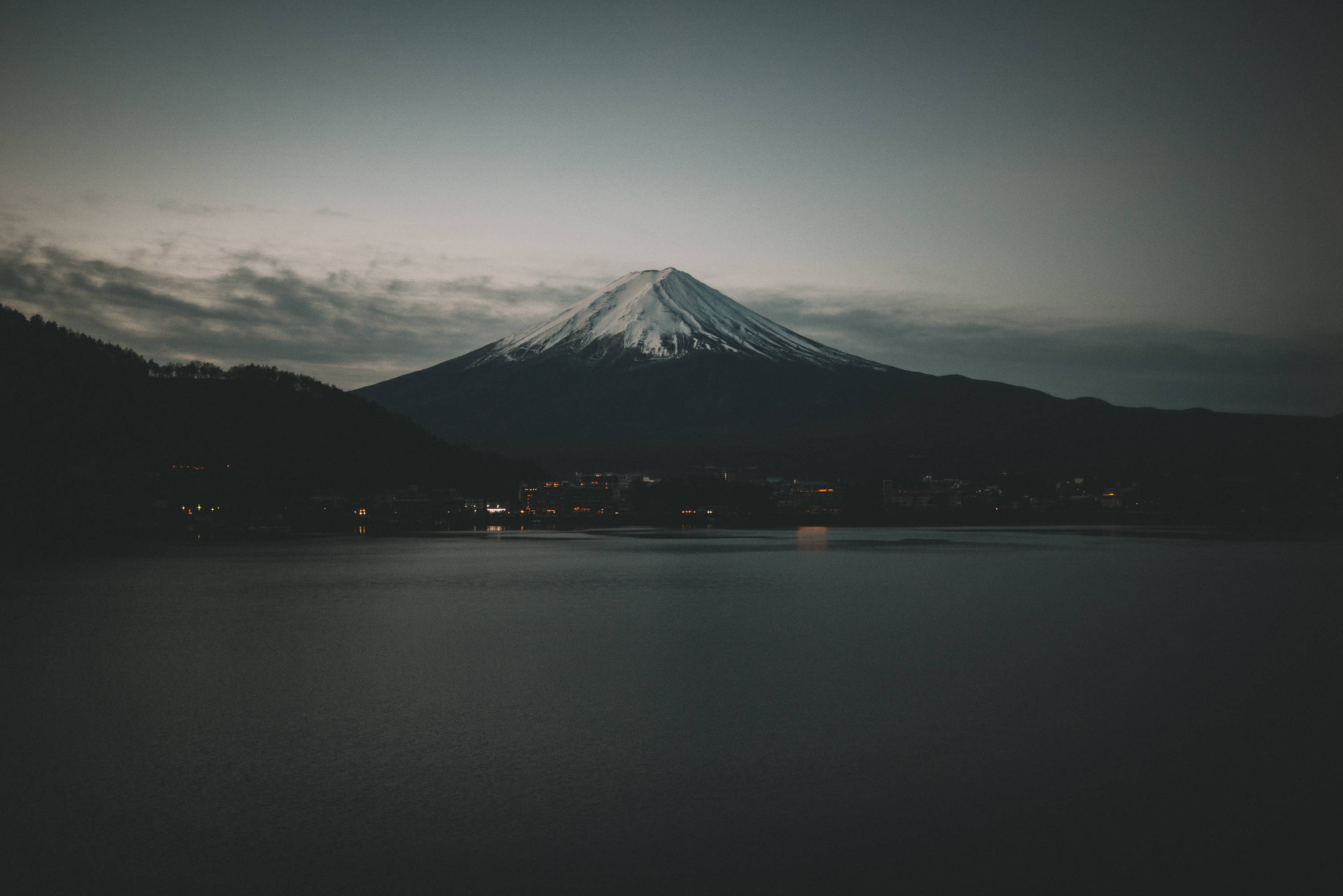 日本 桌布 壁紙 — 白雪覆蓋的富士山在黃昏時分雄偉地聳立在寧靜的湖面上，散落的城鎮燈光倒映在水中。