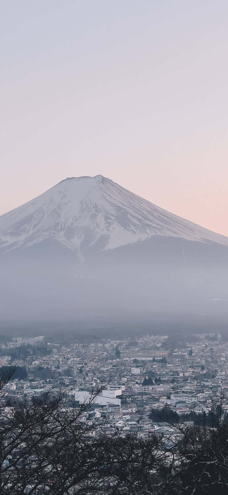 灰色美學動圖 桌布 壁紙 — 白雪覆蓋的富士山在黎明時分雄偉地聳立於霧氣瀰漫的小鎮之上，天空呈現柔和的粉紅色和灰色。