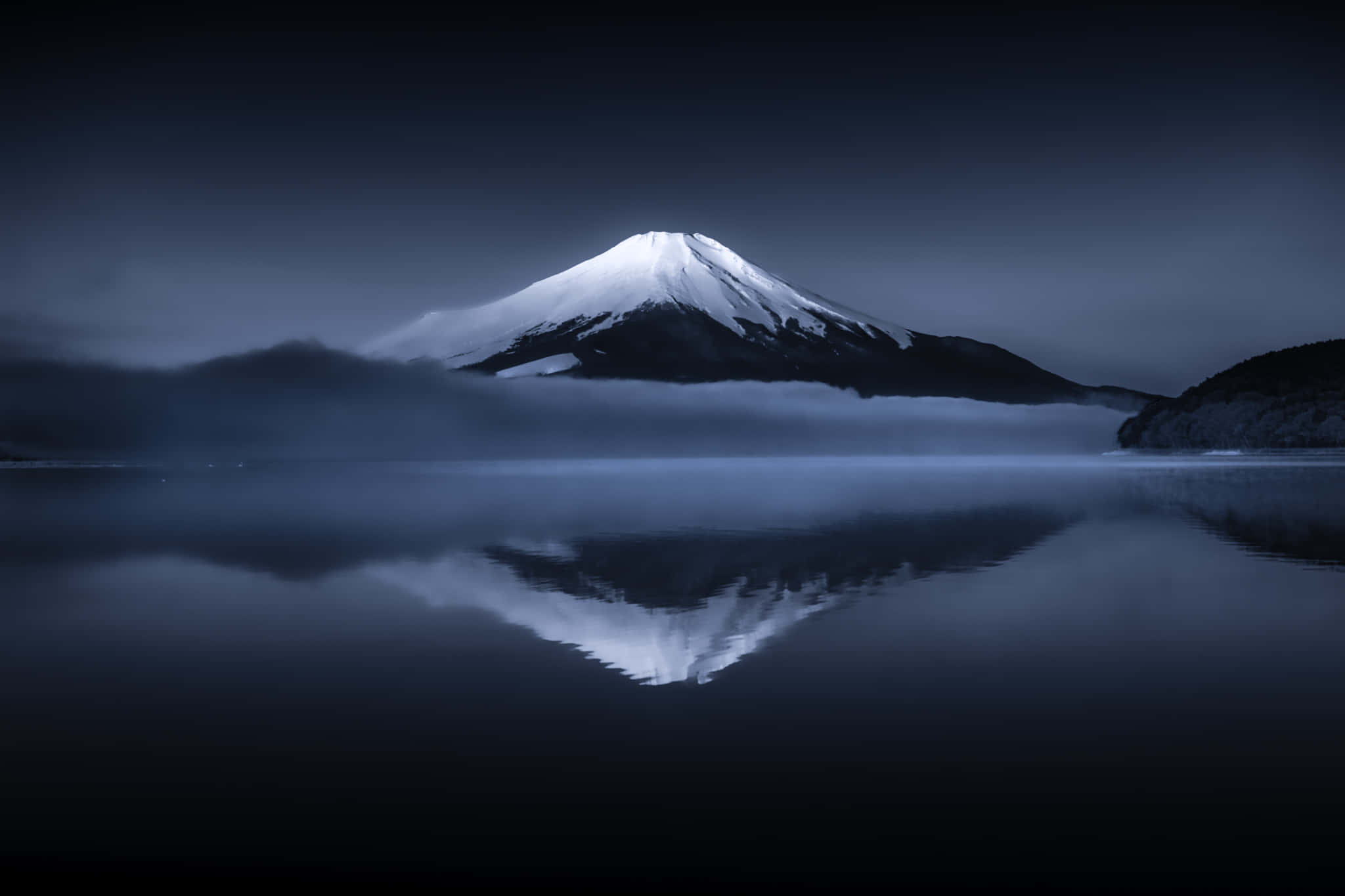 富士山 桌布 壁紙 — 白雪覆蓋的富士山從薄霧中雄偉升起，其完美的倒影映照在玻璃般的湖面上，背景是陰鬱的暮色天空。