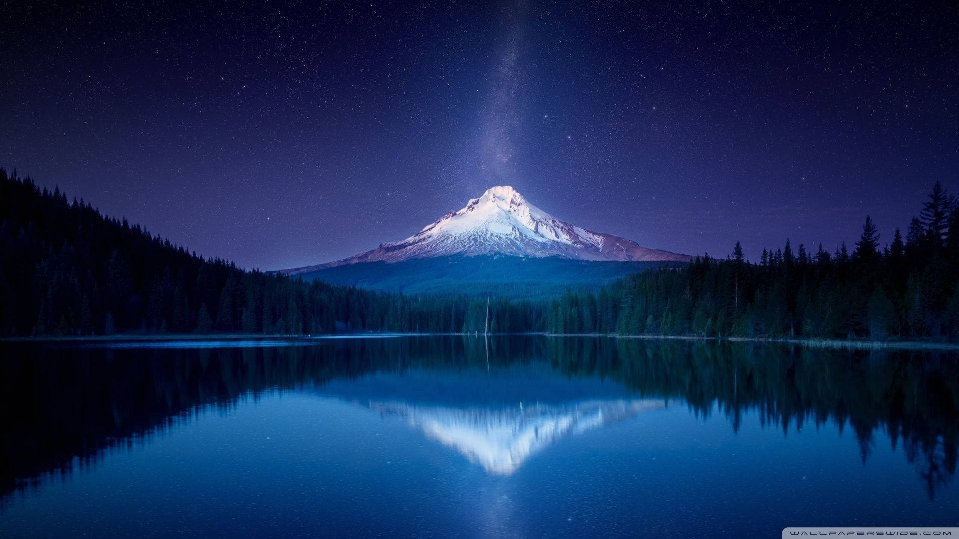 Amazing 桌布 壁紙 — 白雪覆蓋的山峰，在繁星點點的夜空下，可見銀河，完美倒映在寧靜的藍色湖泊中，四周環繞著黑暗的松林。