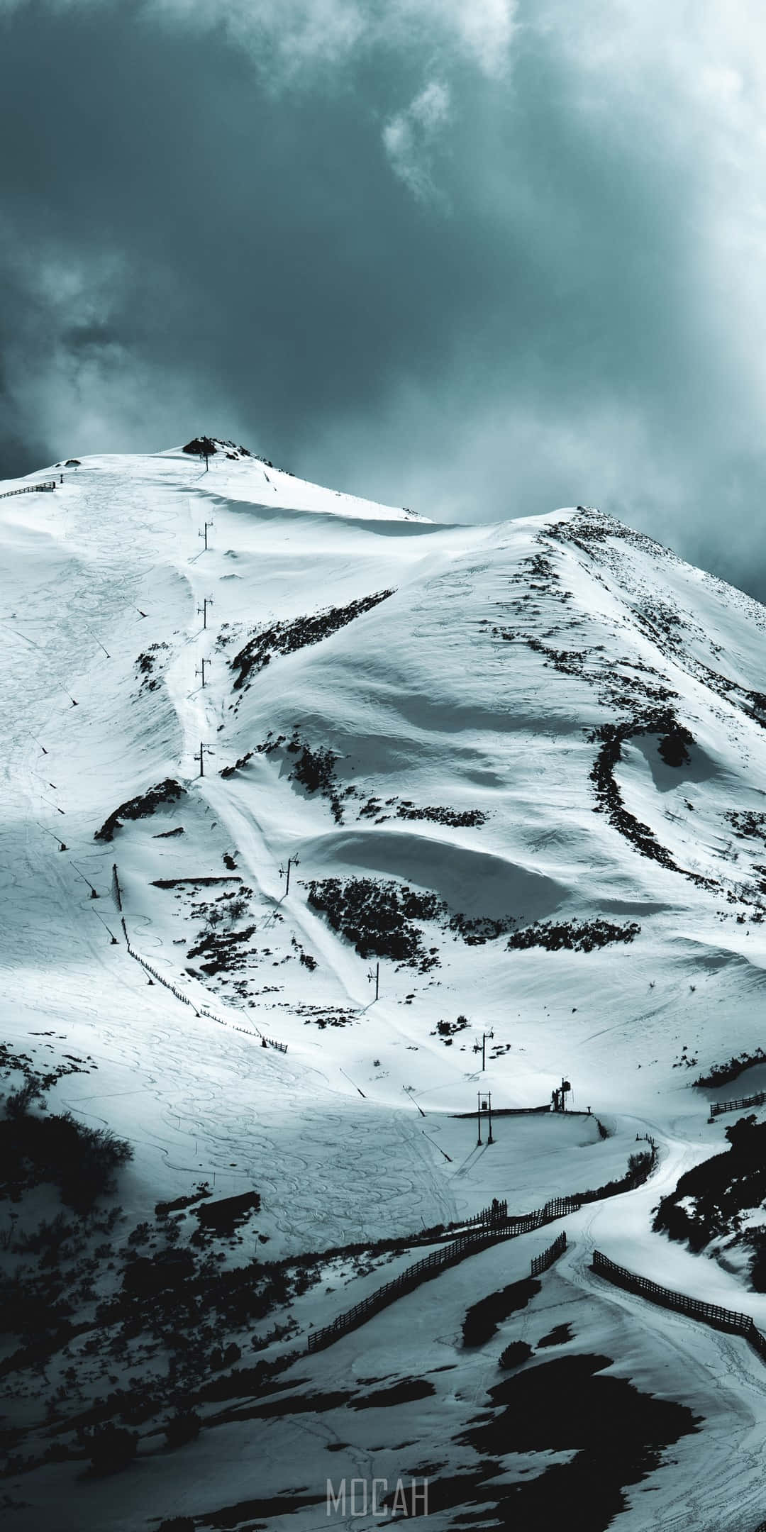 冬天 桌布 壁紙 — 白雪覆蓋的山峰，天空多雲而戲劇化，設有滑雪纜車和蜿蜒的雪道。