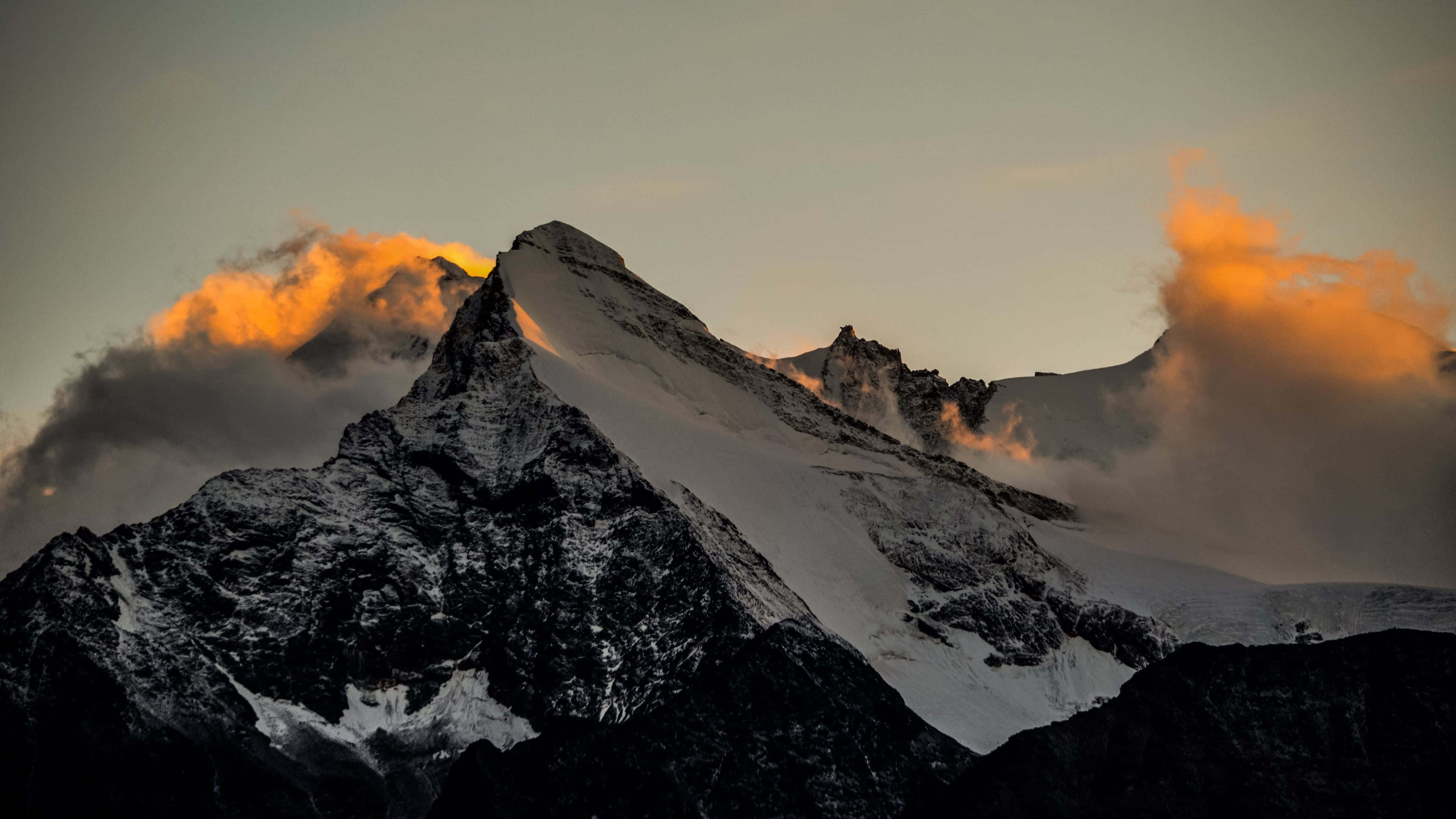macOS 桌布 壁紙 — 白雪覆蓋的山峰刺穿戲劇性的雲層，在日出時發出橙色光芒，下方是黑暗的岩石斜坡。