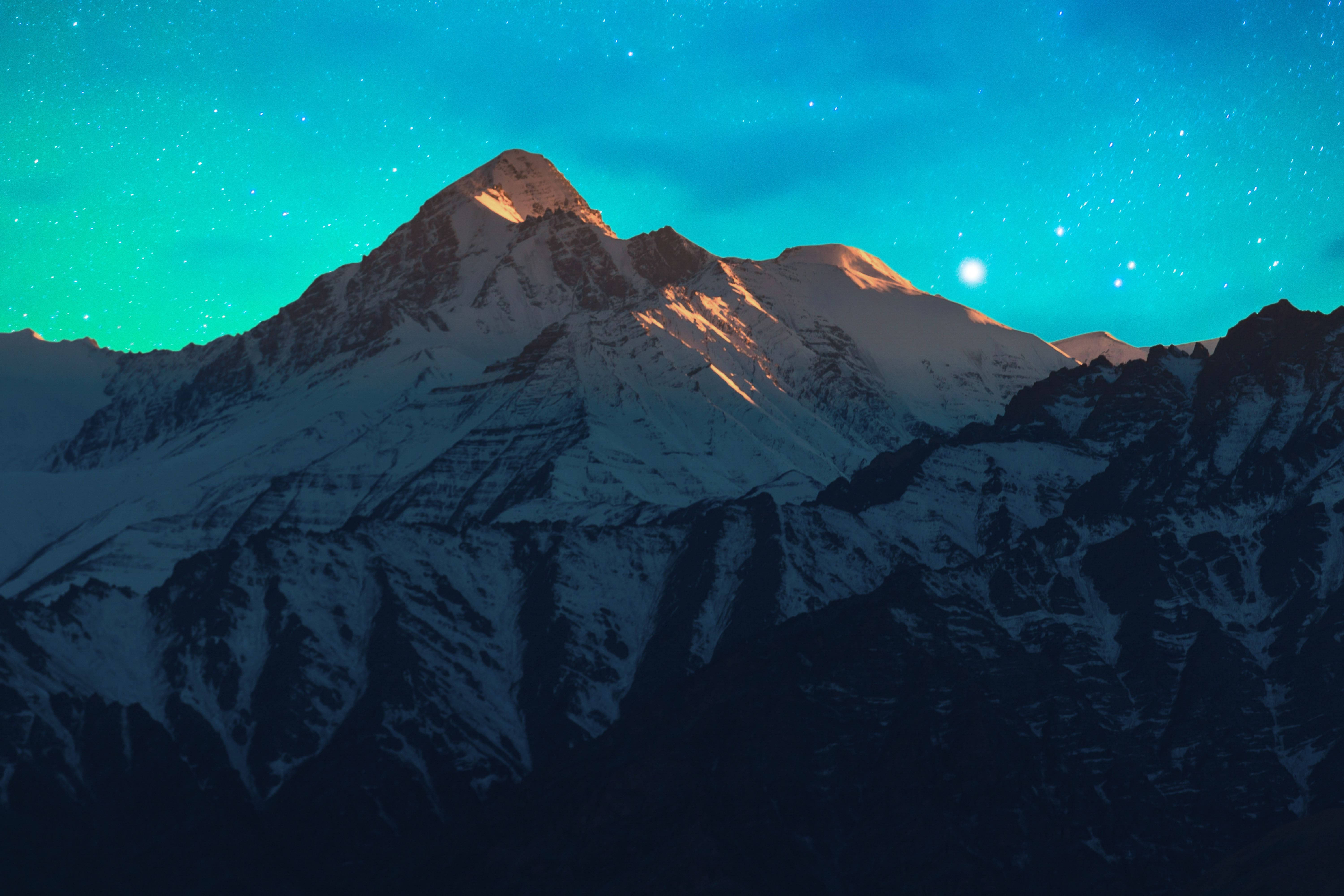 自然 4K 桌布 壁紙 — 白雪覆蓋的山峰在充滿活力的藍綠色星空下閃耀，伴隨著明亮的月亮。