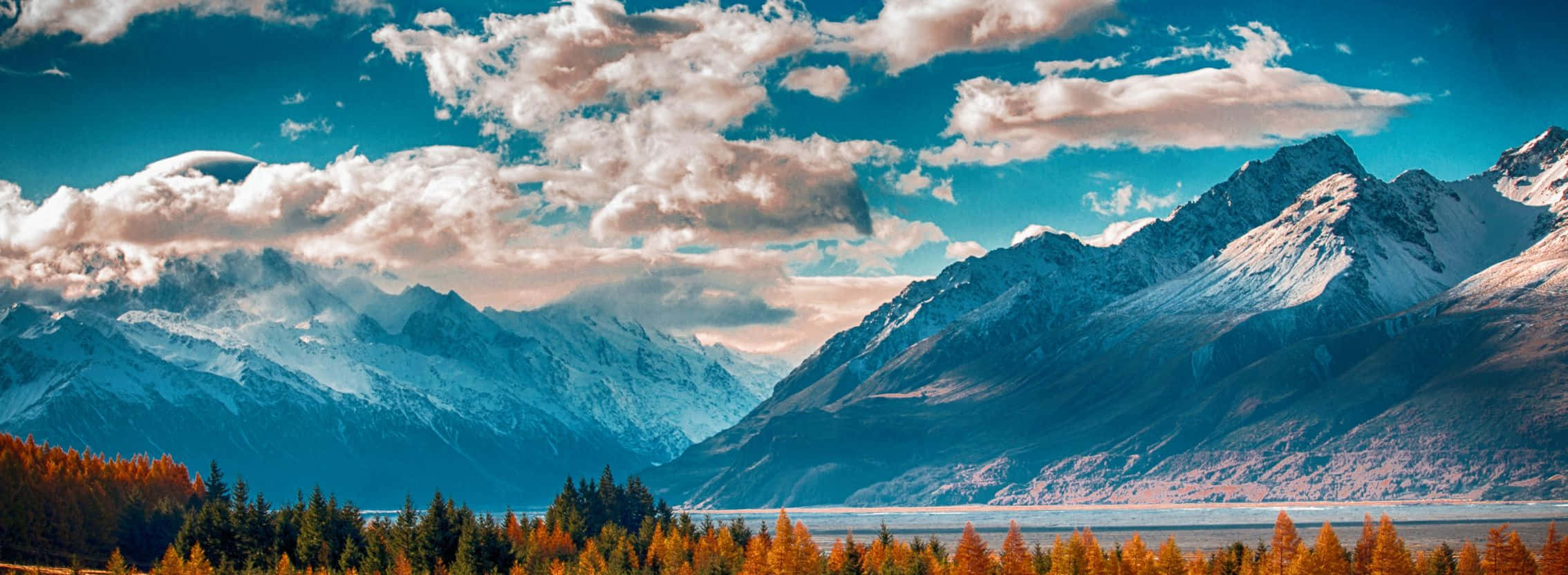 2268x832 桌布 壁紙 — 白雪覆蓋的山峰在寧靜的湖泊後方升起，湖泊被秋日樹木環繞，上方是帶有蓬鬆雲朵的鮮豔藍天。