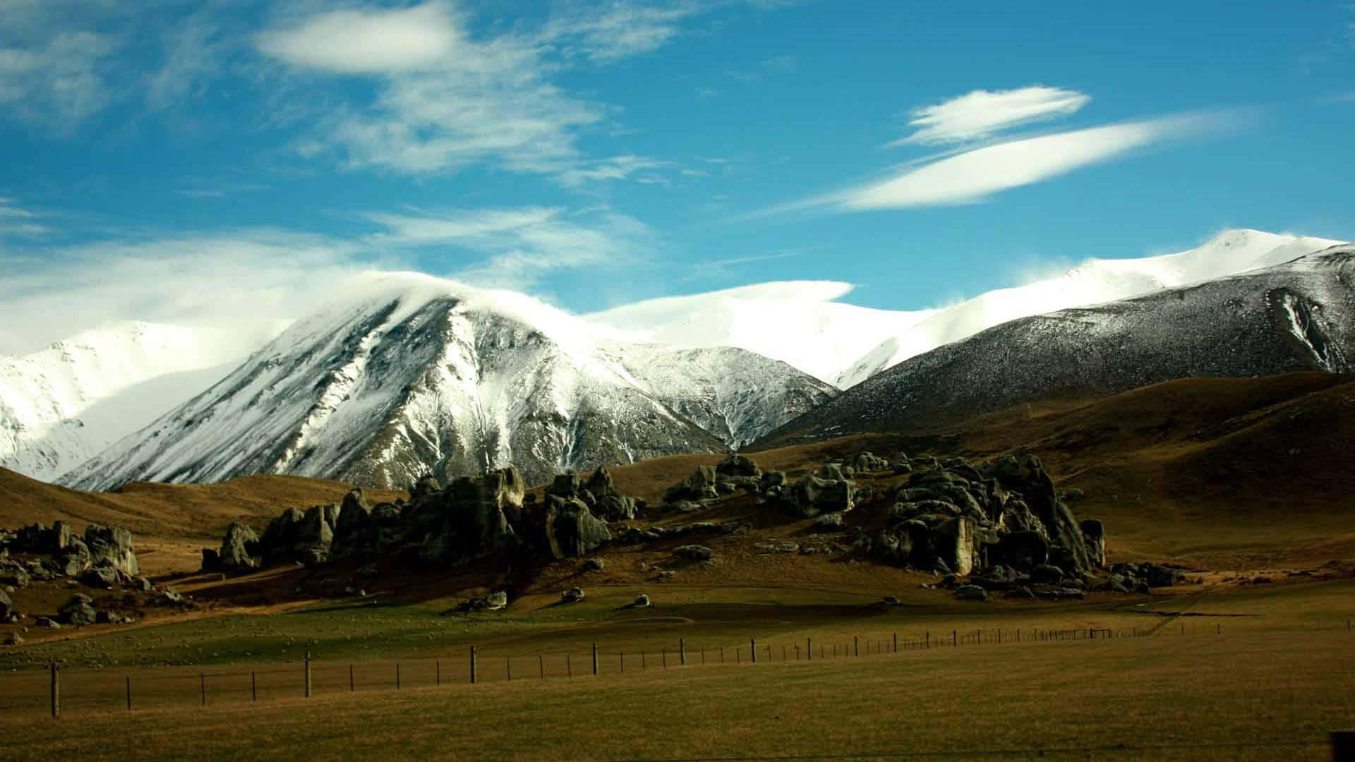 紐西蘭 桌布 壁紙 — 白雪覆蓋的山峰在崎嶇的岩石地帶後方雄偉地聳立，天空湛藍，飄著縷縷輕雲。
