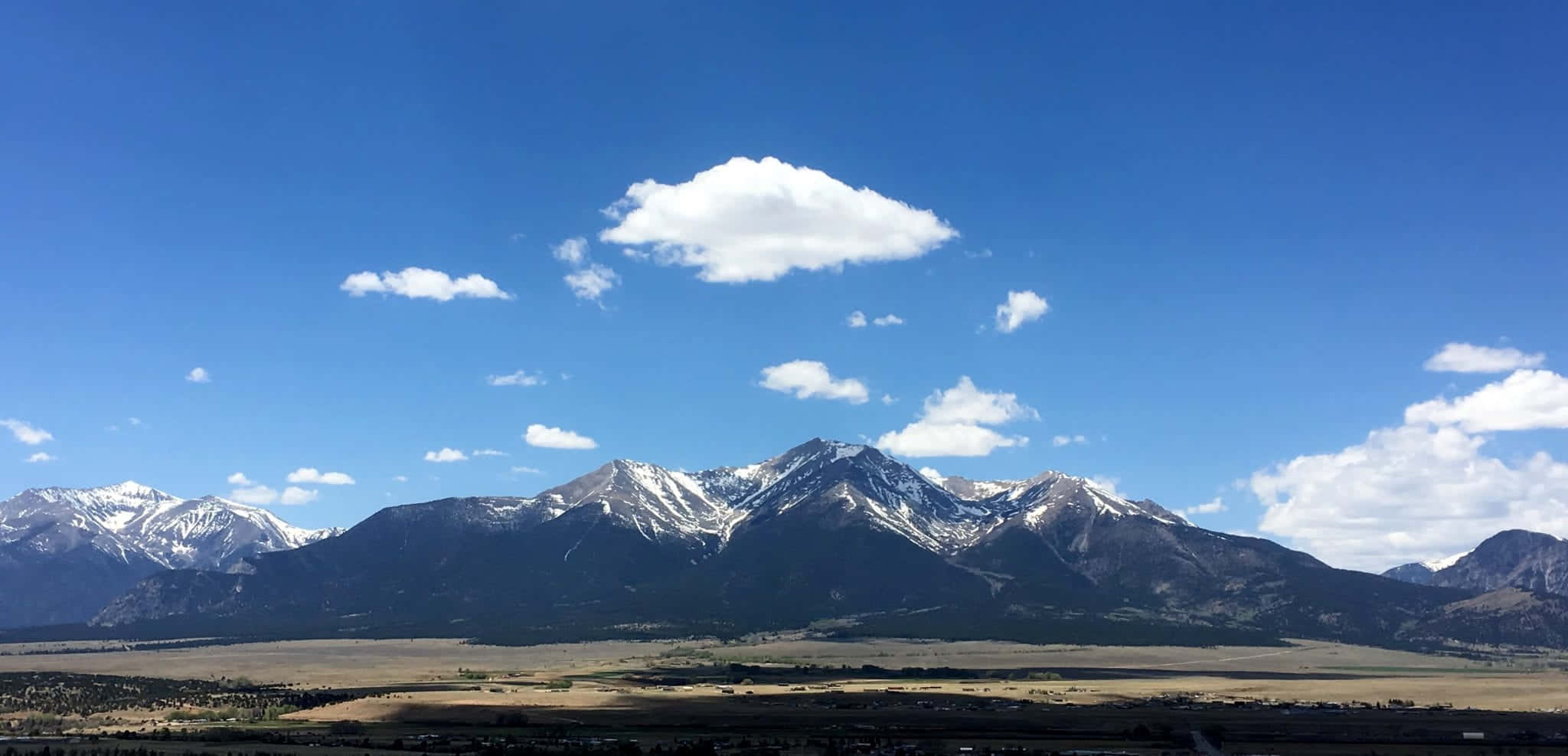 Colorado Mountain 桌布 壁紙 — 白雪覆蓋的山峰在廣闊的藍天下雄偉地聳立，藍天上點綴著蓬鬆的白雲，下方是金色的山谷。