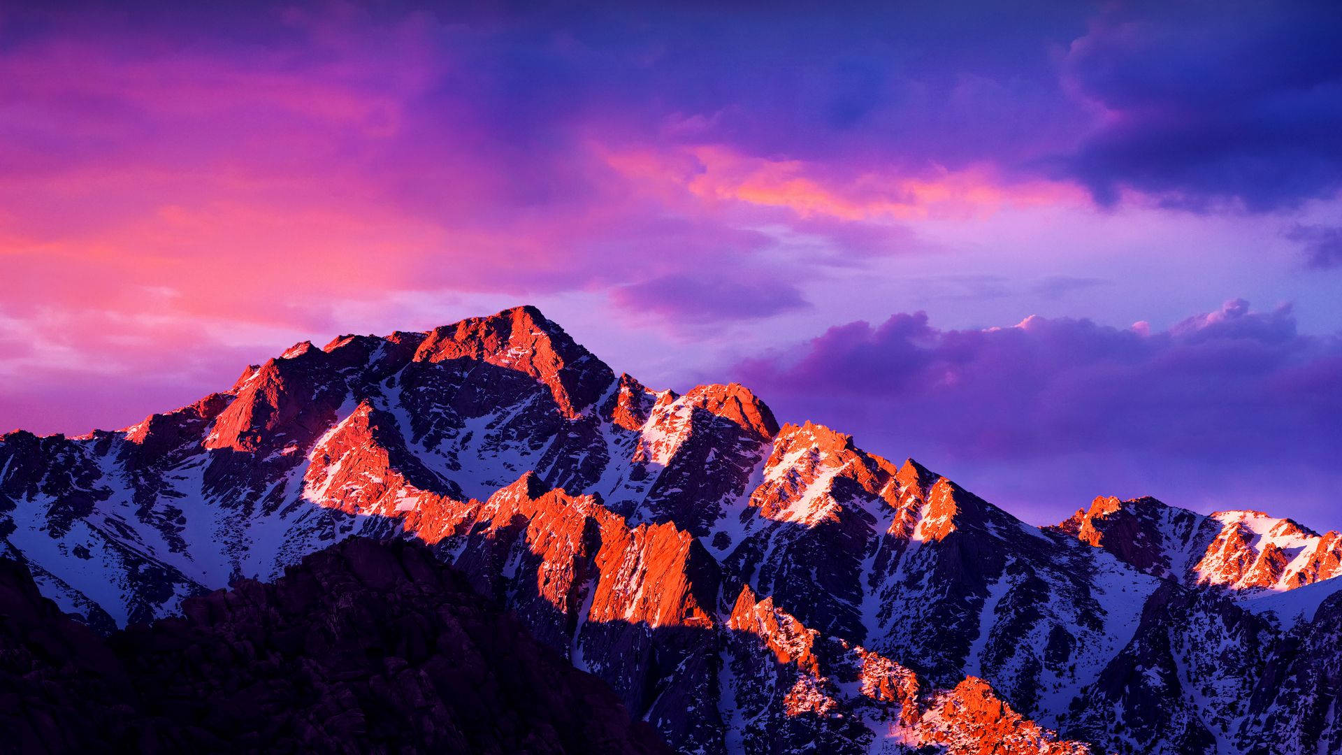 macOS 桌布 壁紙 — 白雪覆蓋的山峰在戲劇性的紫色和粉紅色日落天空映襯下，閃耀著火橙色的光芒。