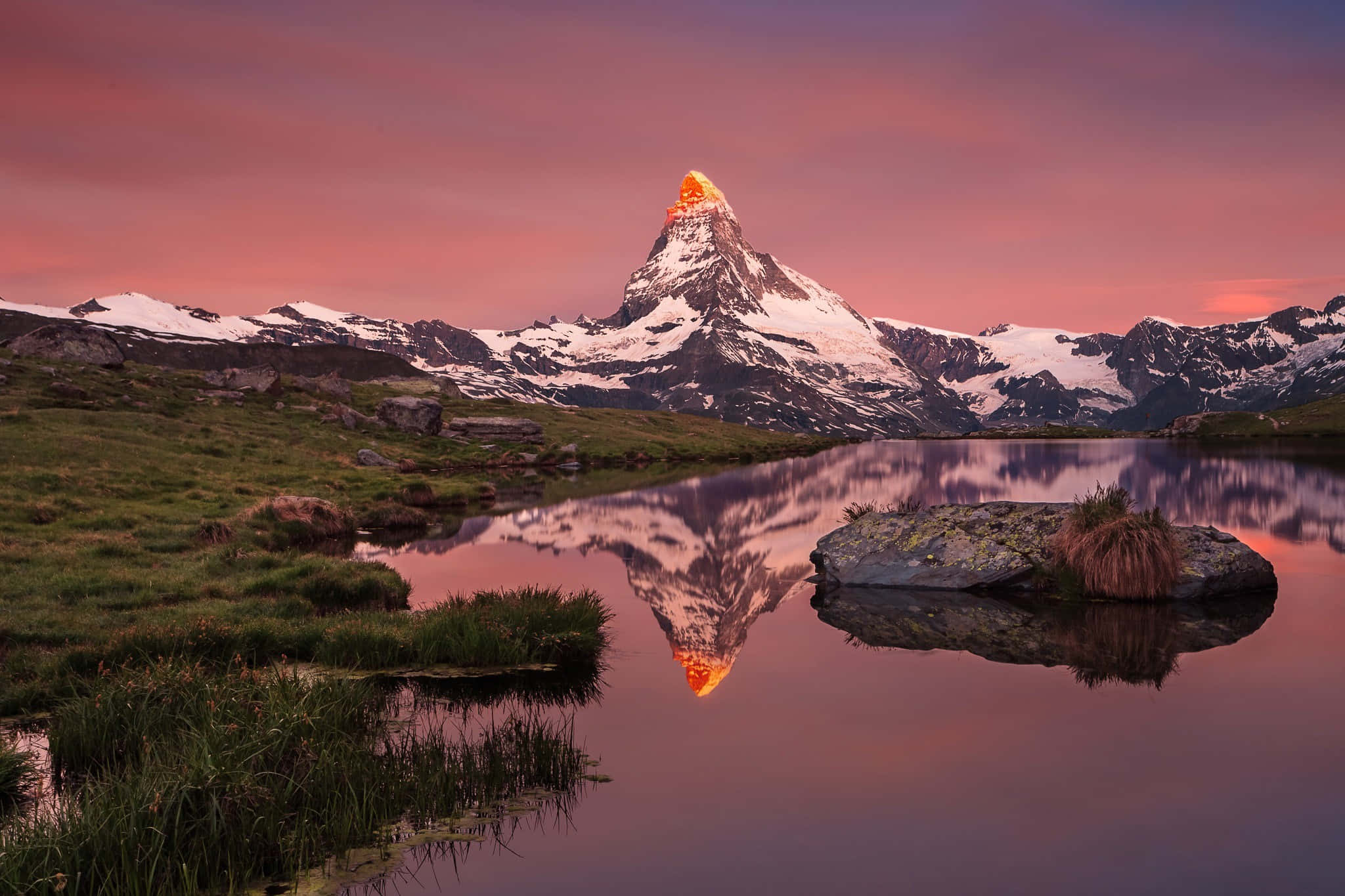 Mountain Scenery 桌布 壁紙 — 白雪覆蓋的山峰在日落時發出橙色光芒，完美倒映在綠草如茵的寧靜高山湖泊中。