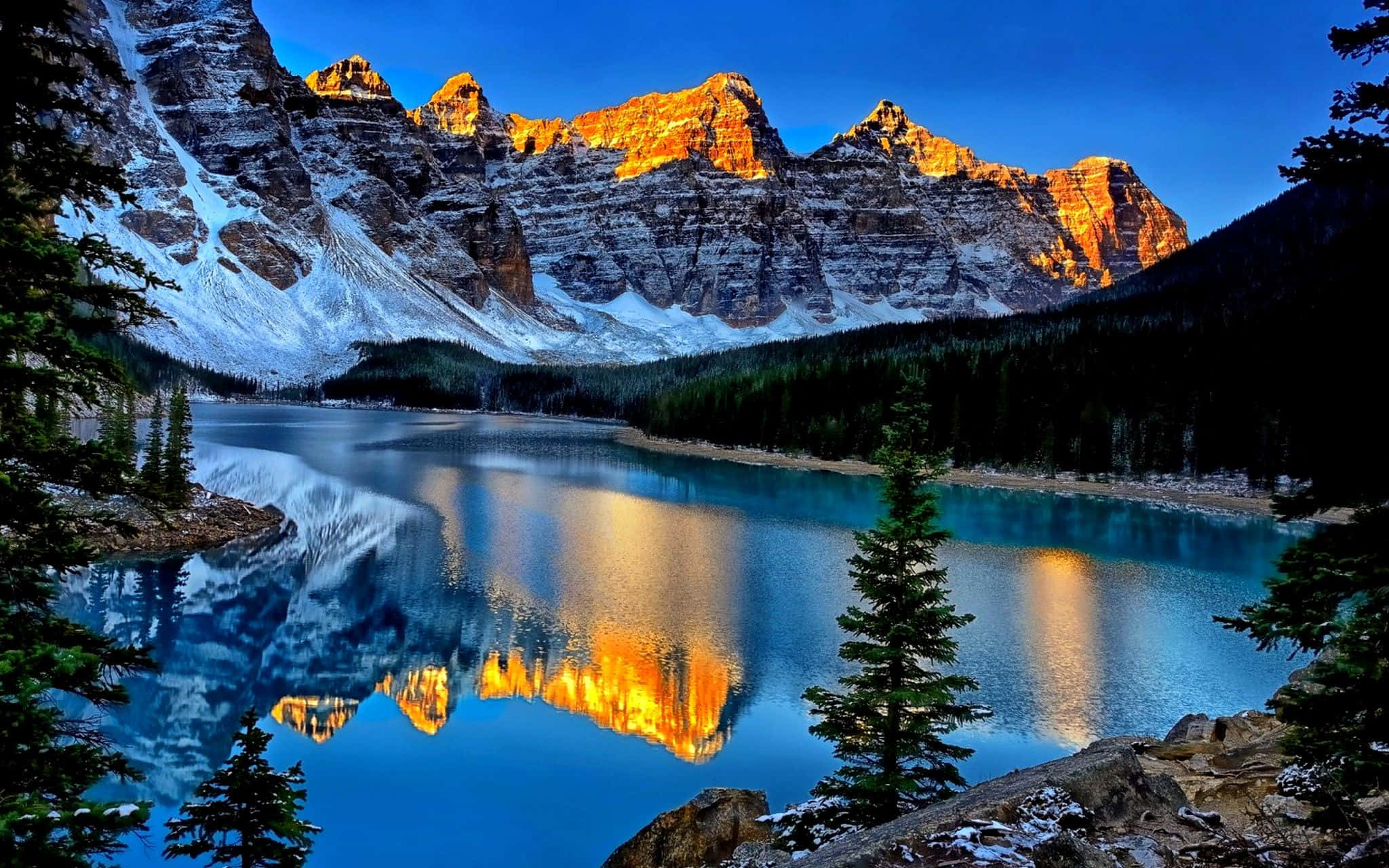 Beautiful Lake 桌布 壁紙 — 白雪覆蓋的山峰在日落時閃耀金光，完美倒映在由常綠樹環繞的寧靜碧綠湖泊中。