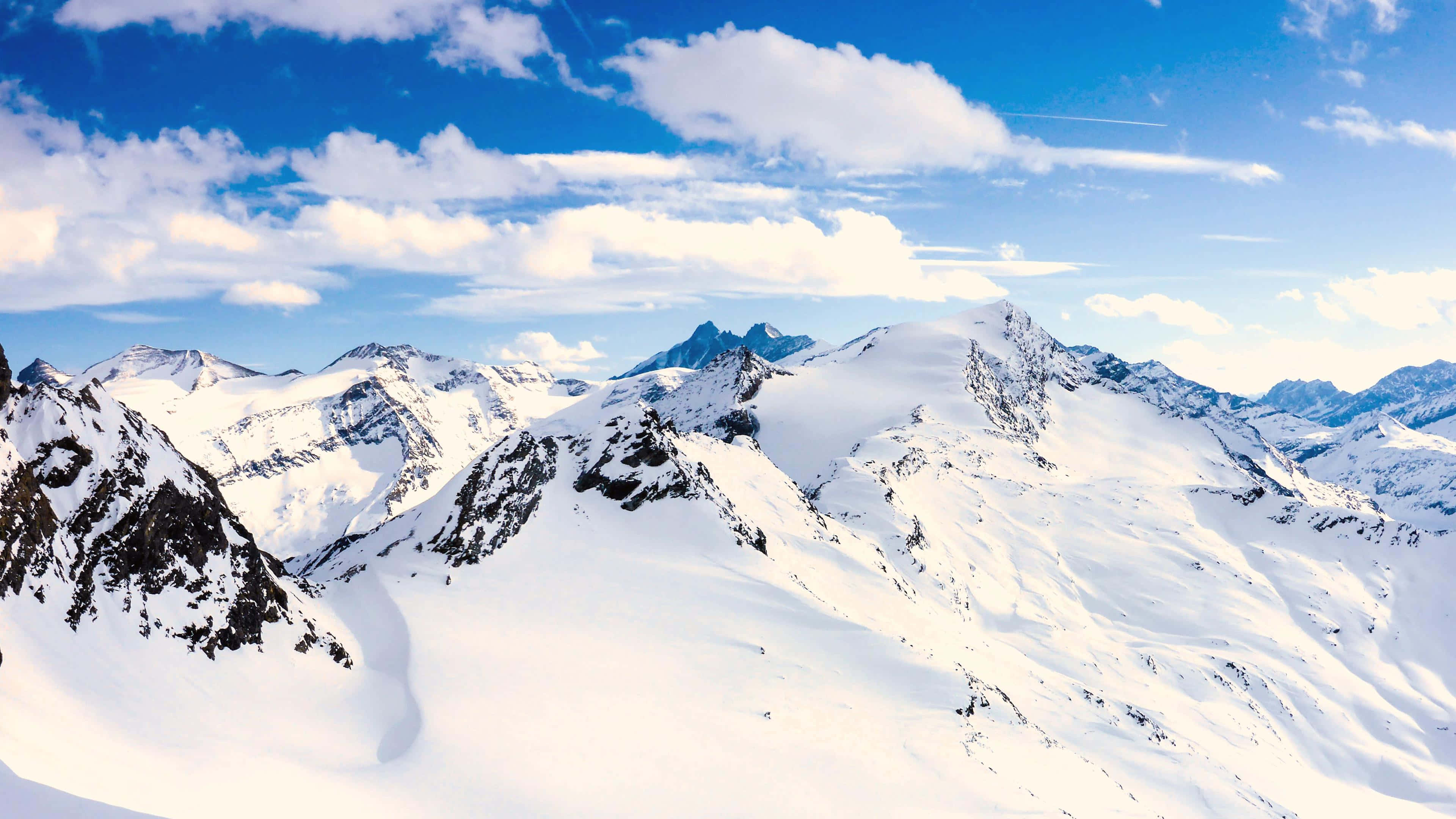4K Mountain 桌布 壁紙 — 白雪覆蓋的山峰在明亮的日光下刺穿點綴著蓬鬆白雲的鮮豔藍天。