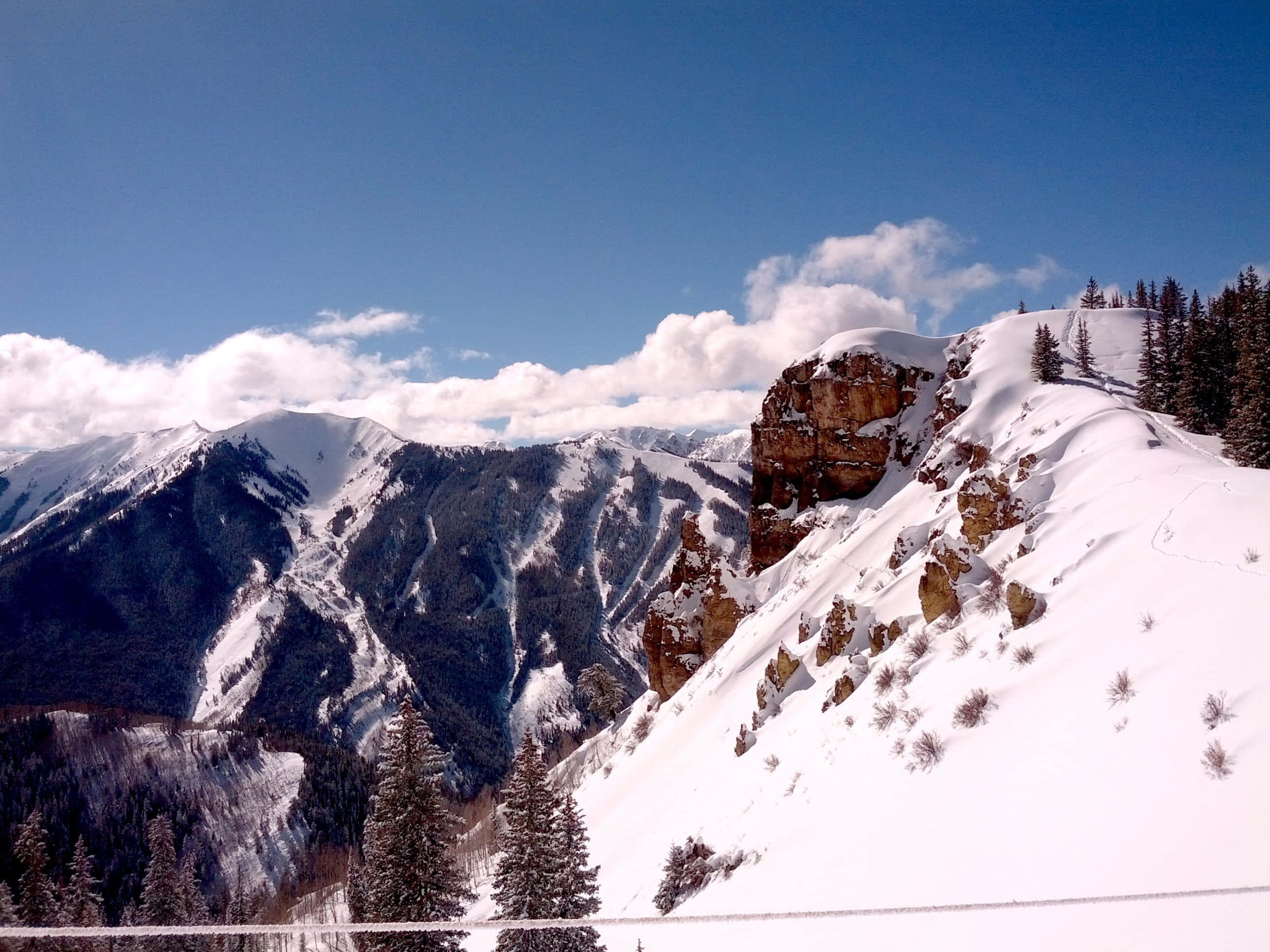 Aspen Colorado 桌布 壁紙 — 白雪覆蓋的山峰在明亮的藍天下聳立，蓬鬆的白雲和常綠樹點綴著山坡。