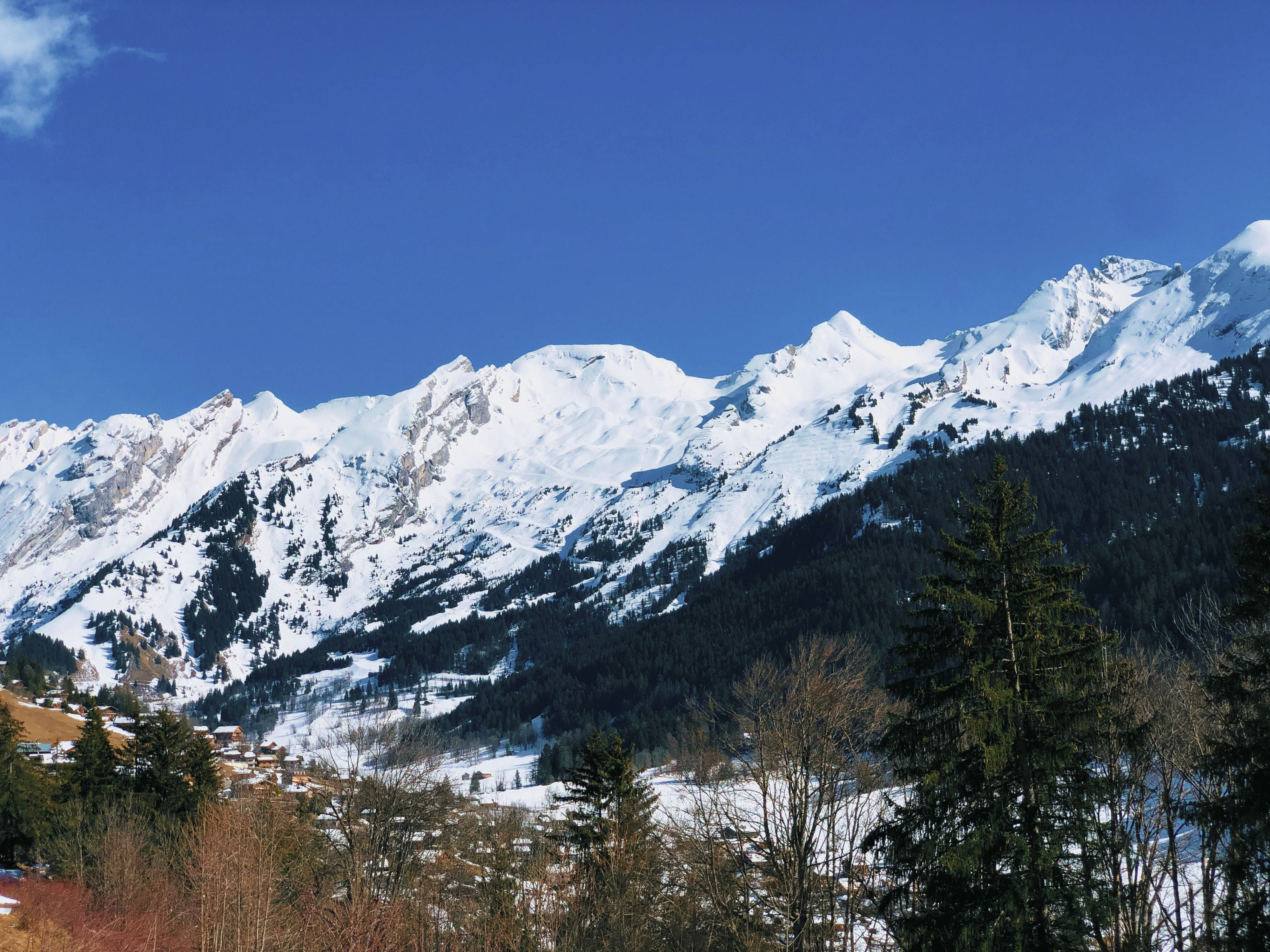 Calming 桌布 壁紙 — 白雪覆蓋的山峰在晴朗的藍天下，聳立於森林覆蓋的山谷之上，山谷中散落著房屋。