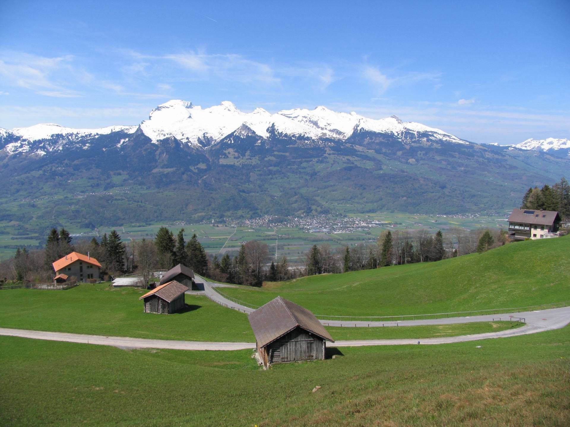 Liechtenstein 桌布 壁紙 — 白雪覆蓋的山峰在晴朗藍天下，從散佈著房屋和蜿蜒道路的山谷後方升起。