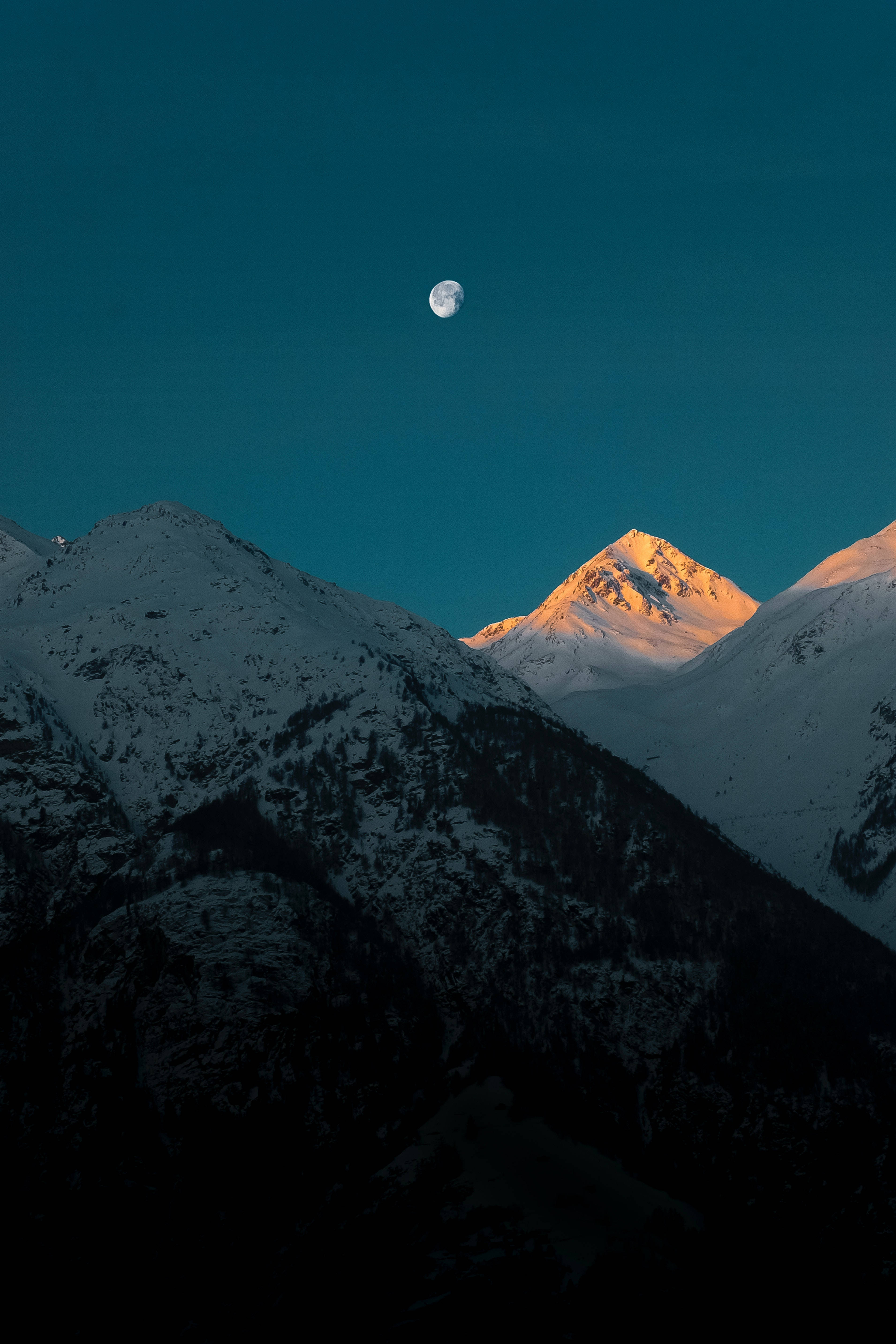 Blue Sky 桌布 壁紙 — 白雪覆蓋的山峰在溫暖的光芒中閃耀，月亮懸掛在深青色的暮色天空中。