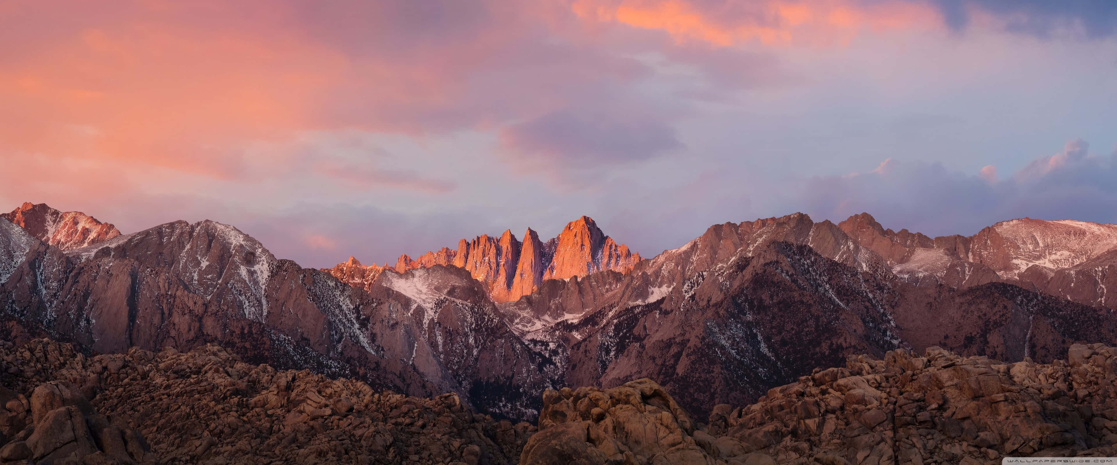 Apple 3840x1600 桌布 壁紙 — 白雪覆蓋的山峰在溫暖的粉紅色和橙色色調中閃耀，夕陽落在崎嶇的岩石山脊後面。