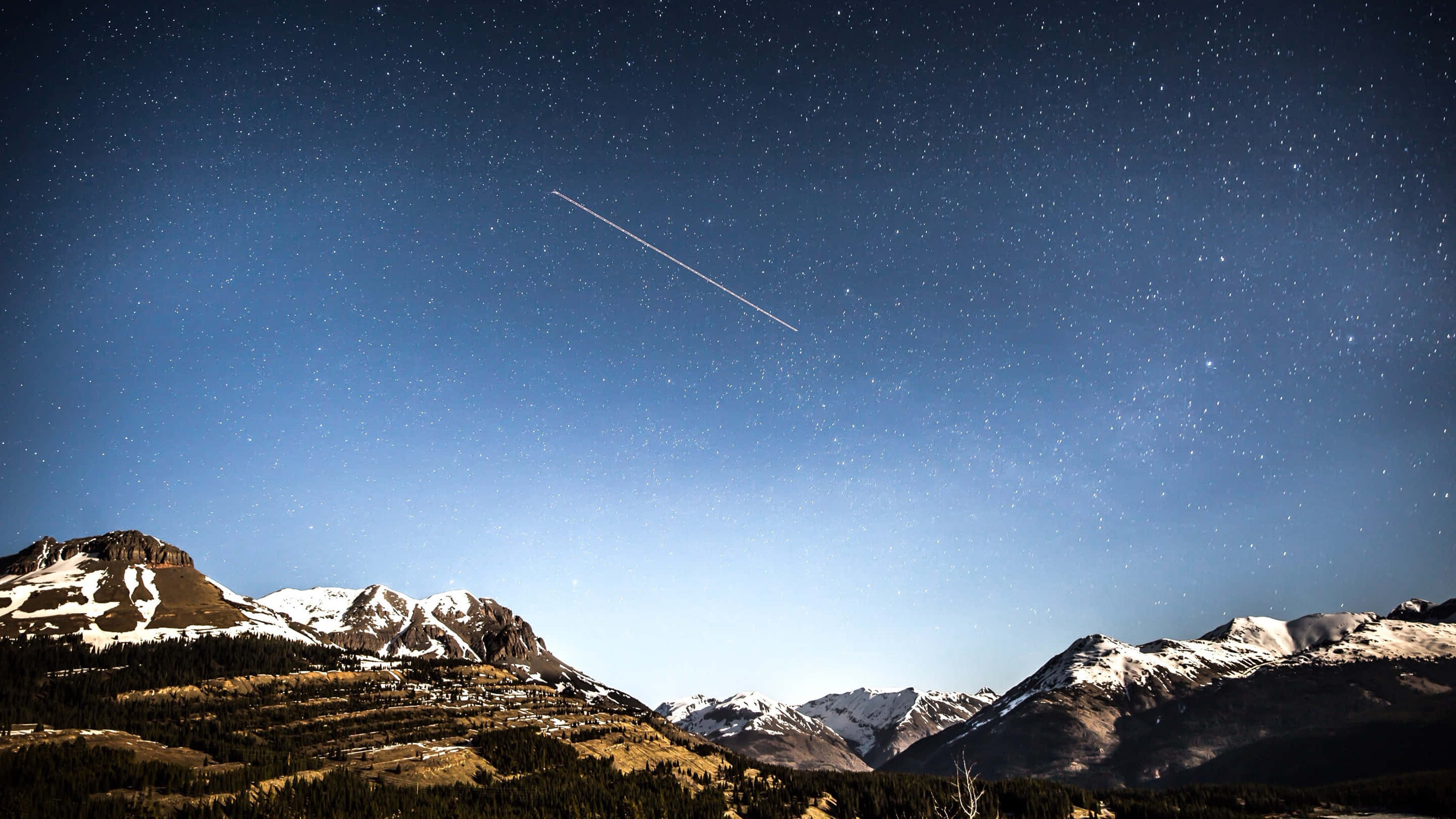 三星 桌布 壁紙 — 白雪覆蓋的山峰在繁星點點的夜空下聳立，一道明亮的飛機尾跡劃過天際。