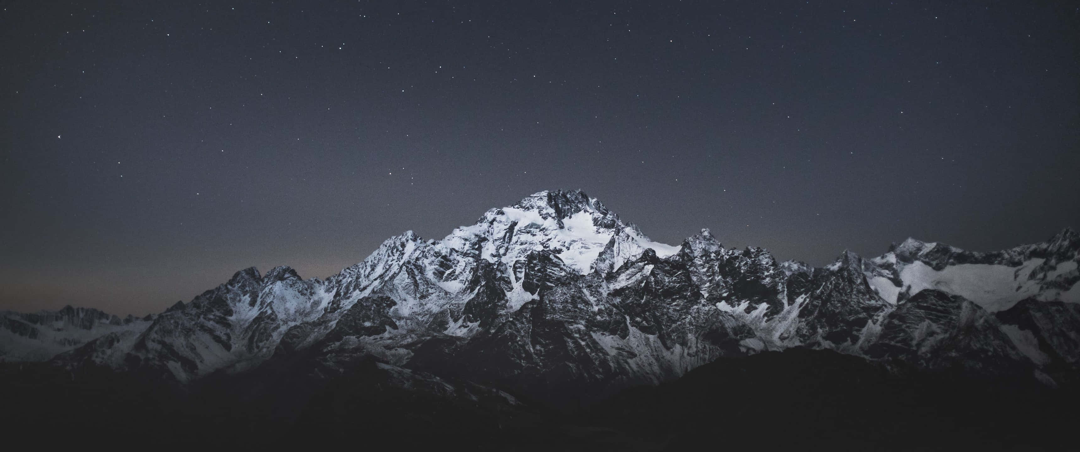Winter 桌布 壁紙 — 白雪覆蓋的山峰在繁星點點的夜空和地平線上微弱的極光映襯下，巍然聳立。