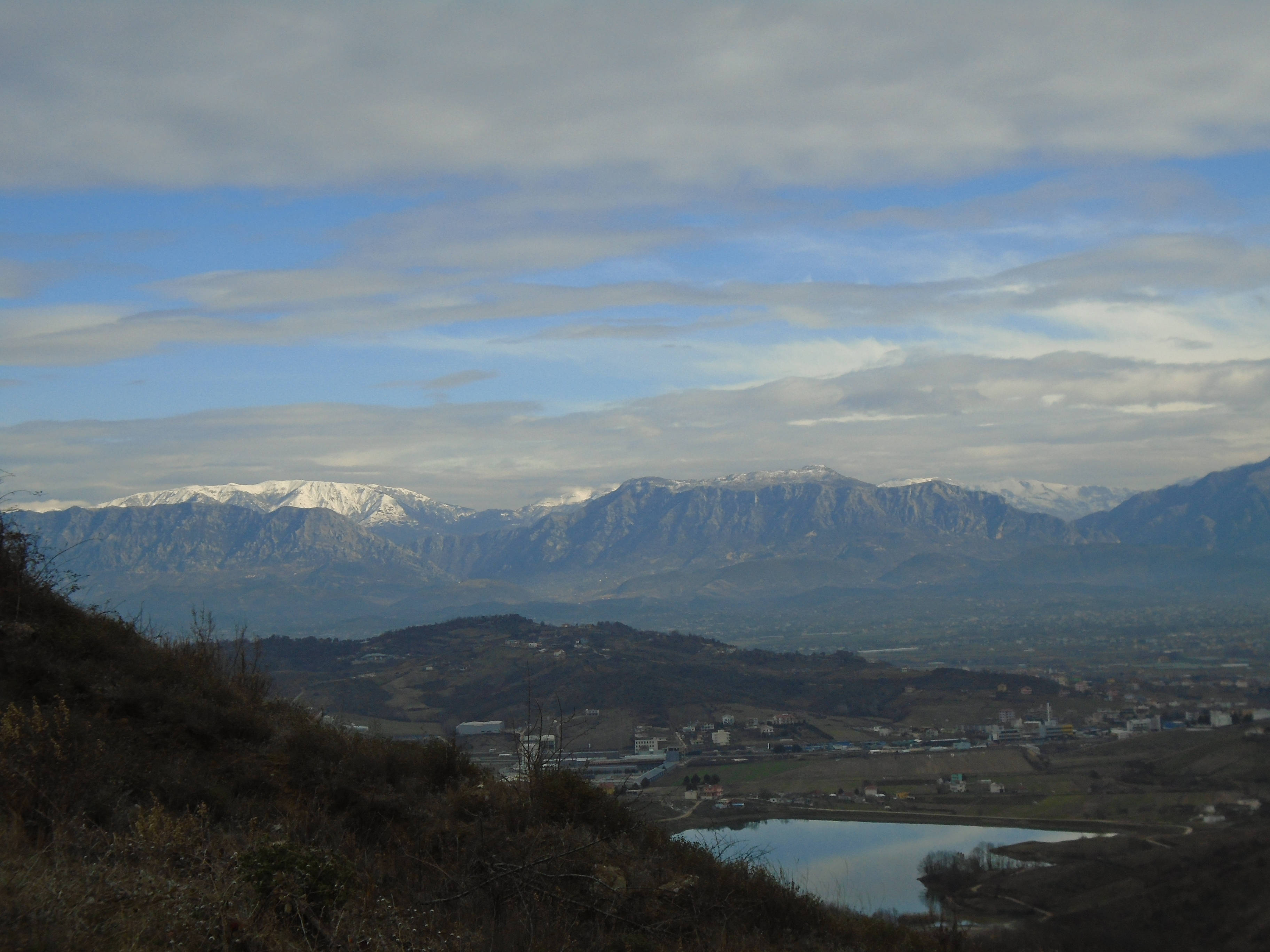 Albania 桌布 壁紙 — 白雪覆蓋的山峰在谷城後方升起，平靜的湖泊倒映著多雲的天空。