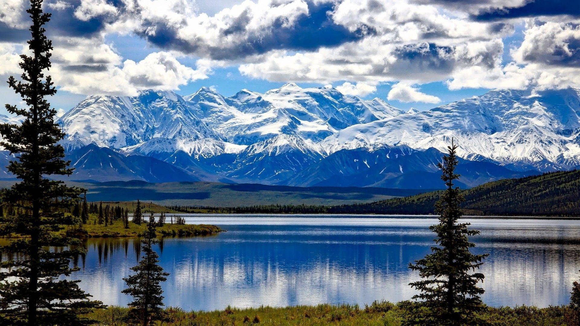 Lake 桌布 壁紙 — 白雪覆蓋的山峰在部分多雲的天空下，在常綠樹環繞的平靜藍色湖泊後方雄偉地升起。