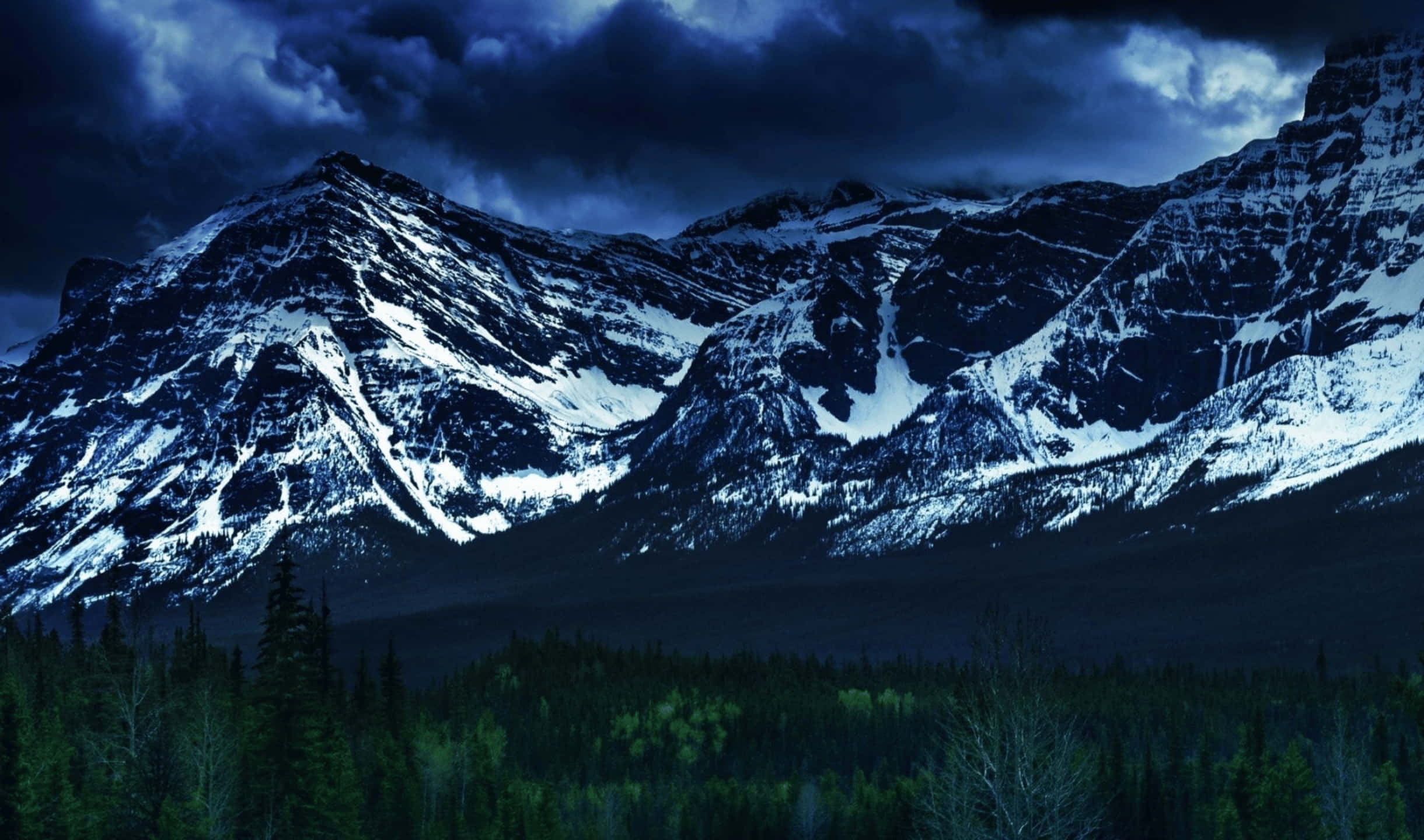 2440x1440 Monitor 桌布 壁紙 — 白雪覆蓋的山峰在風暴暮色天空下，聳立於茂密的常綠森林之上。