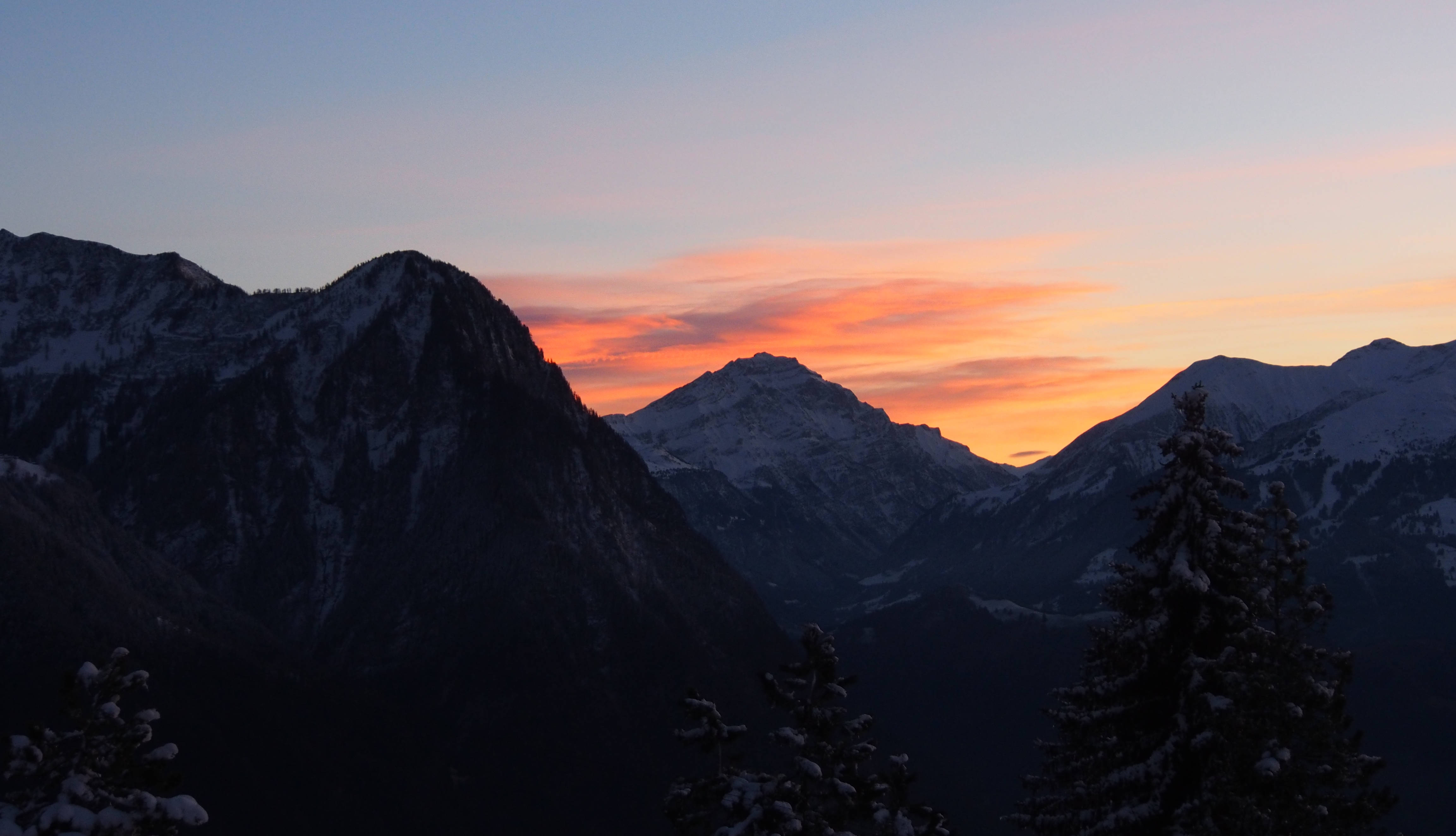 Liechtenstein 桌布 壁紙 — 白雪覆蓋的山峰在鮮豔的橙色和粉紅色日落天空的襯托下形成剪影，前景有深色松樹。