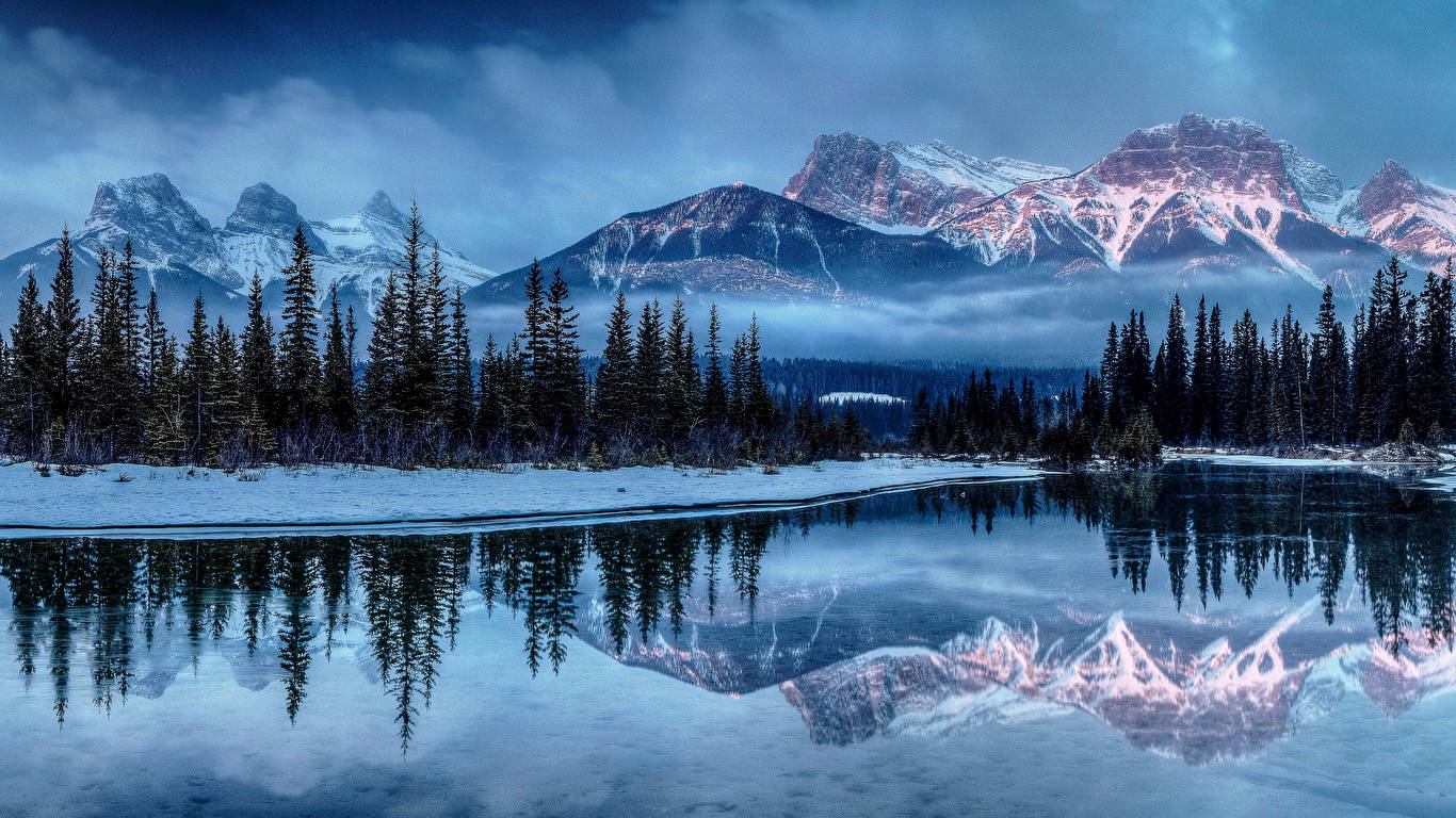 山景 桌布 壁紙 — 白雪覆蓋的山峰在黎明時分閃耀著粉紅色和紫色，完美地倒映在一個被常綠樹環繞的玻璃般清澈的湖泊中。