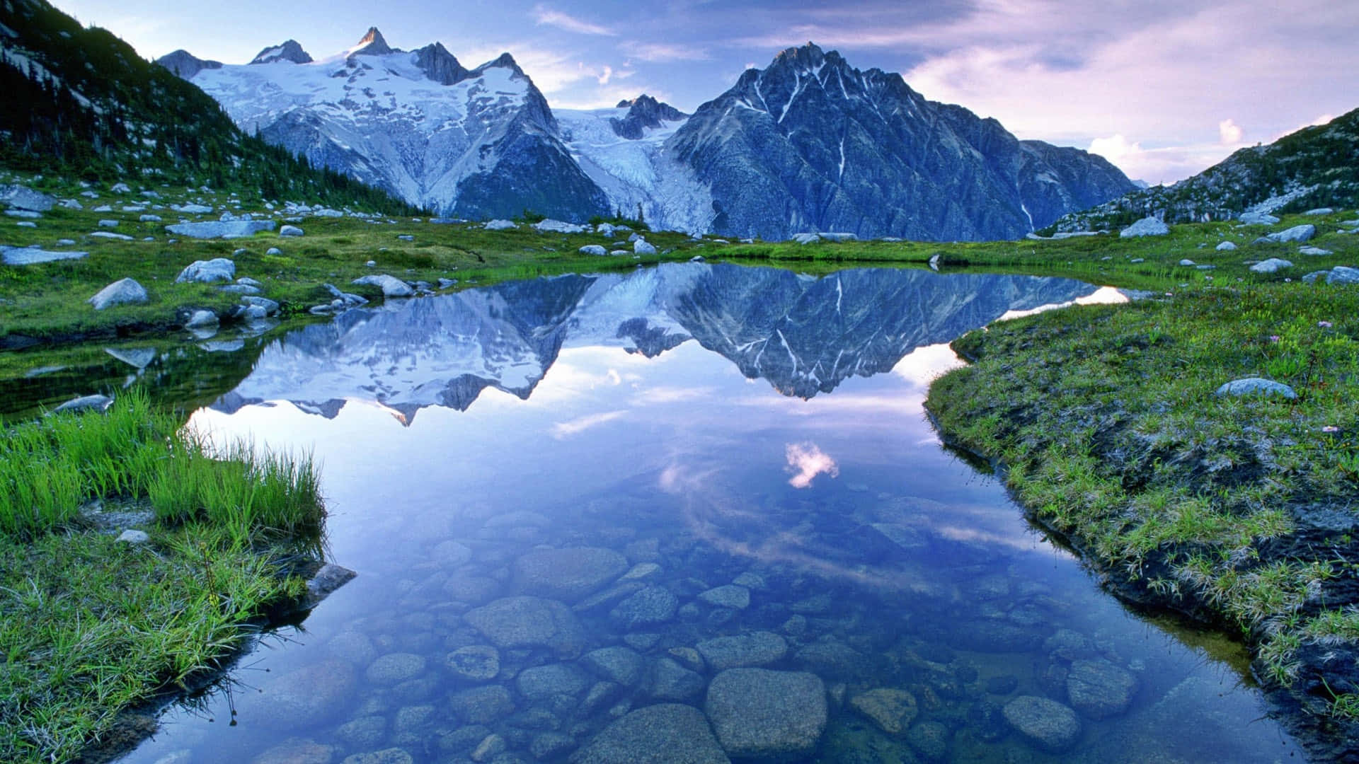 Lake 桌布 壁紙 — 白雪覆蓋的山峰完美地倒映在寧靜的高山湖泊中，周圍是綠色的草地和散落的岩石。