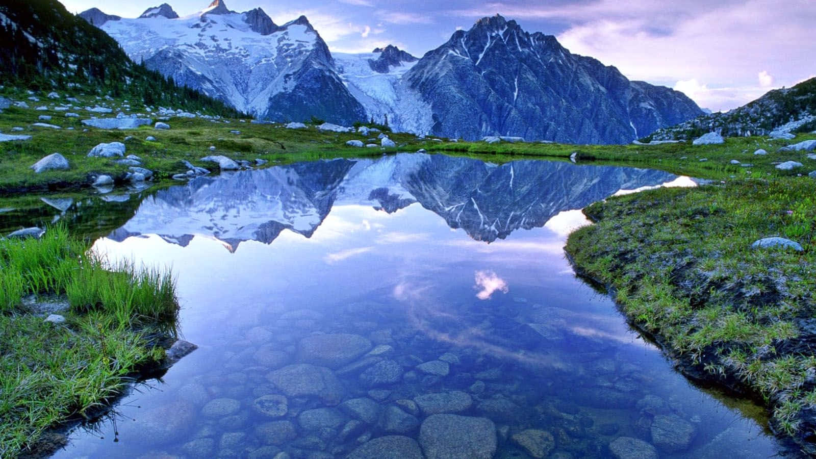Beautiful Lake 桌布 壁紙 — 白雪覆蓋的山峰完美地倒映在寧靜的高山湖泊中，四周環繞著翠綠的草地和散落的岩石。