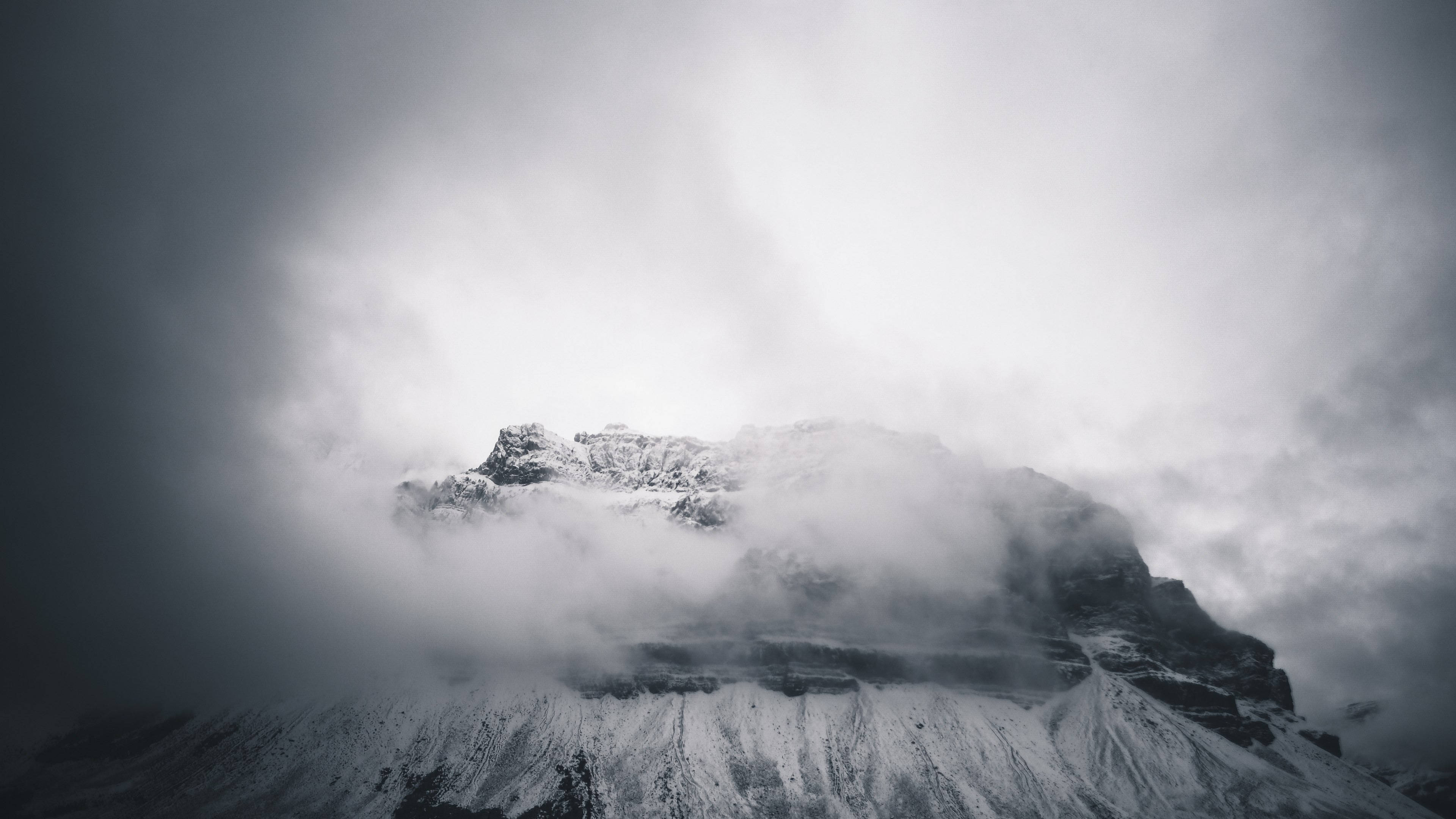 風景 桌布 壁紙 — 白雪覆蓋的山峰從翻滾的雲層中浮現，背景是陰沉的灰色天空。