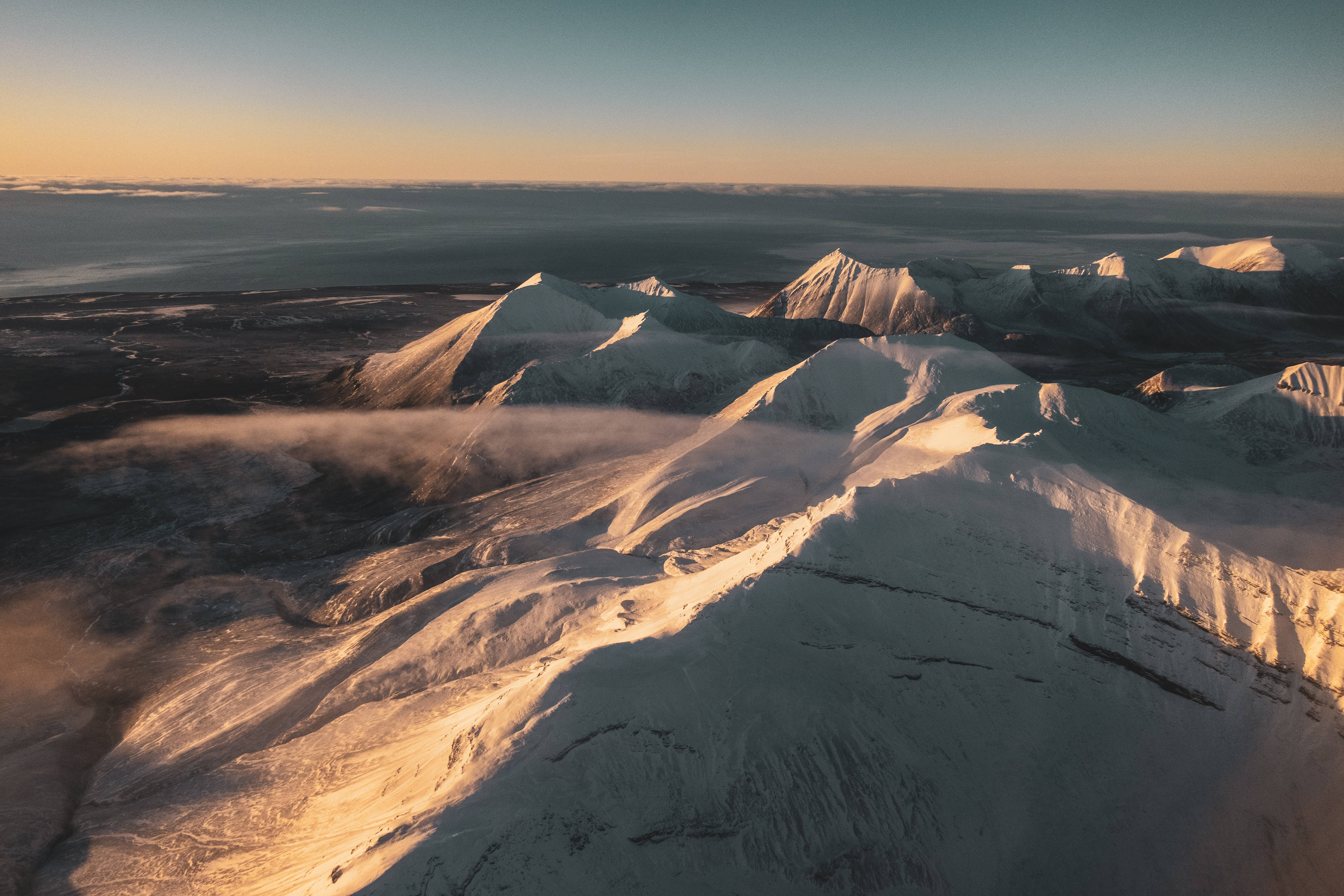 Svalbard 桌布 壁紙 — 白雪覆蓋的山峰沐浴在金色日出光芒中，下方薄霧繚繞的鳥瞰圖。