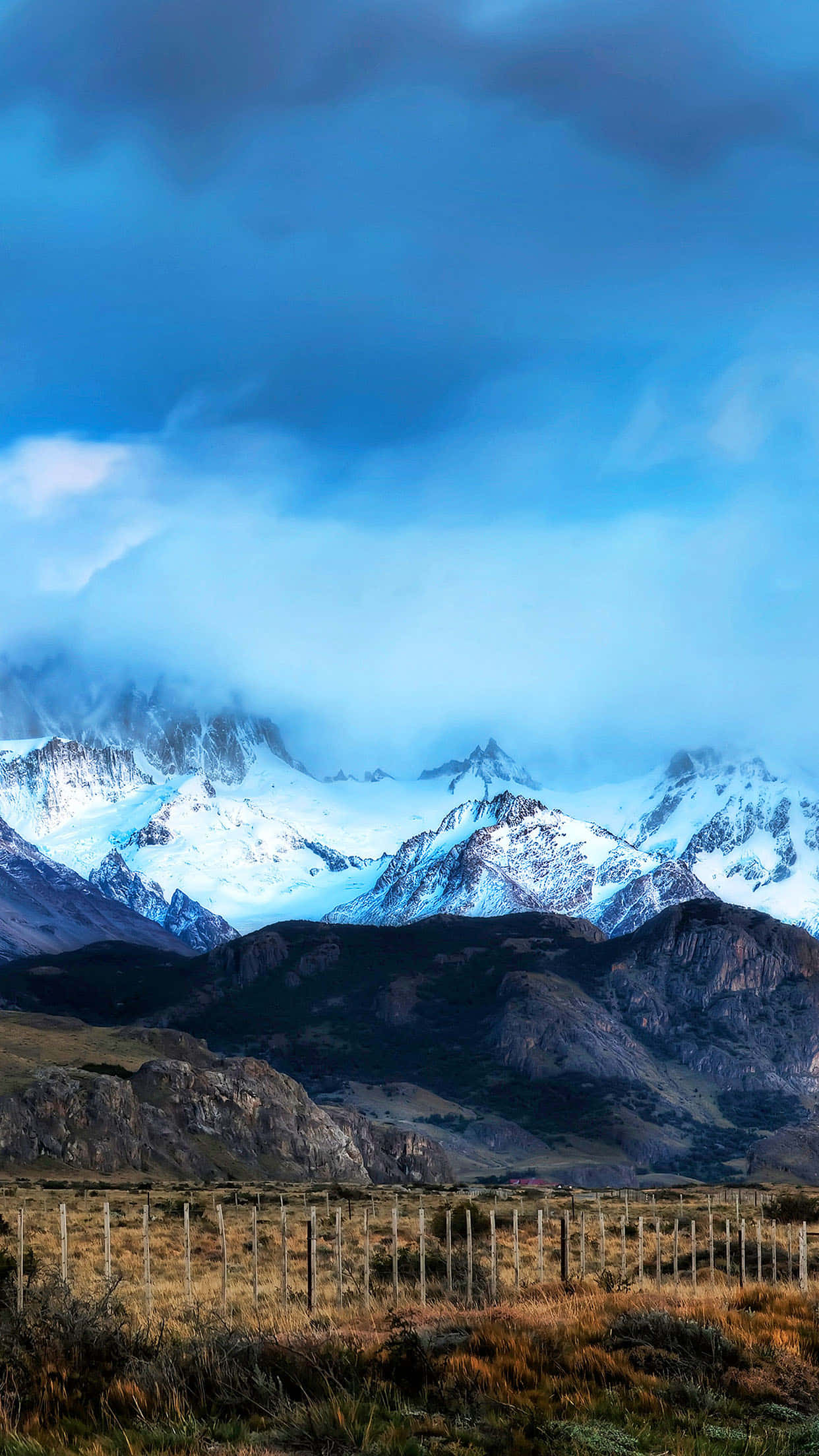 Ranch 桌布 壁紙 — 白雪覆蓋的山峰穿過戲劇性的藍色雲層，下方是崎嶇的山谷，有乾草和木柵欄。