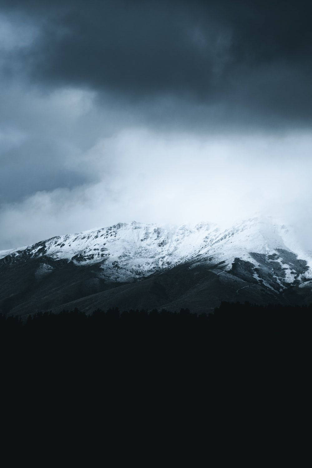 直式 桌布 壁紙 — 白雪覆蓋的山峰穿過黑暗的暴風雲，下方是茂密的森林輪廓。