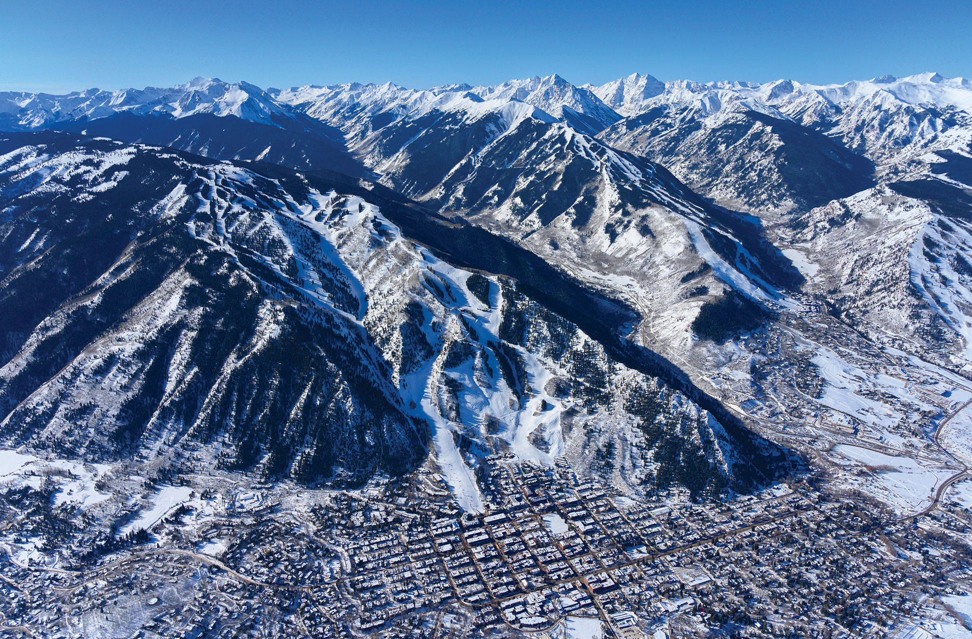Colorado 桌布 壁紙 — 白雪覆蓋的山峰鳥瞰圖，滑雪道在清澈藍天下延伸至冰封的山谷小鎮。