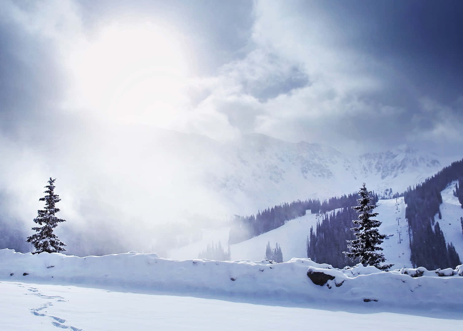 冬季 桌布 壁紙 — 白雪覆蓋的山景，常青樹在明亮的陽光穿透雲層下。