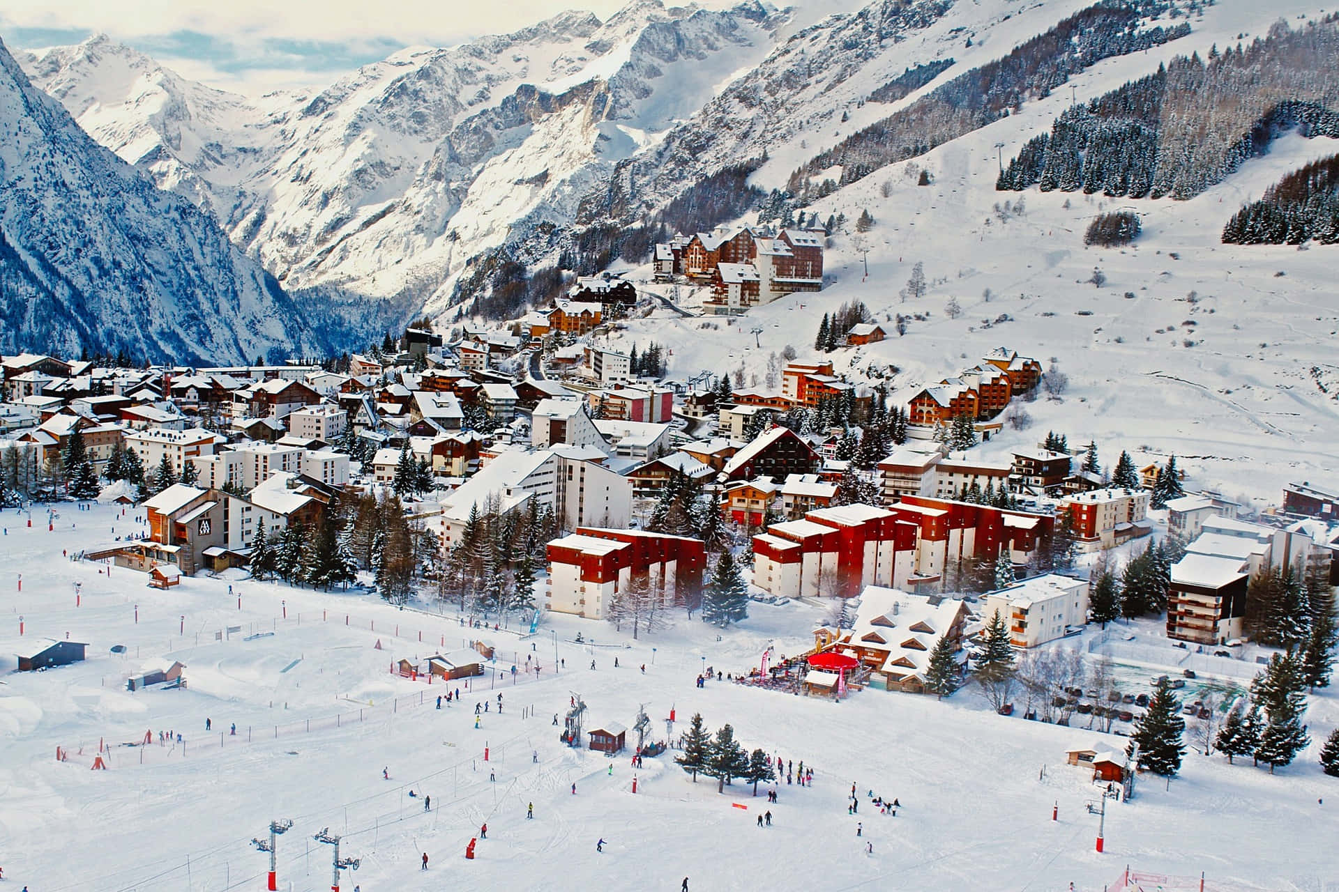 Ski Resort 桌布 壁紙 — 白雪覆蓋的山村鳥瞰圖，紅白相間的小木屋隱藏在松樹和擠滿滑雪者的滑雪道之間。
