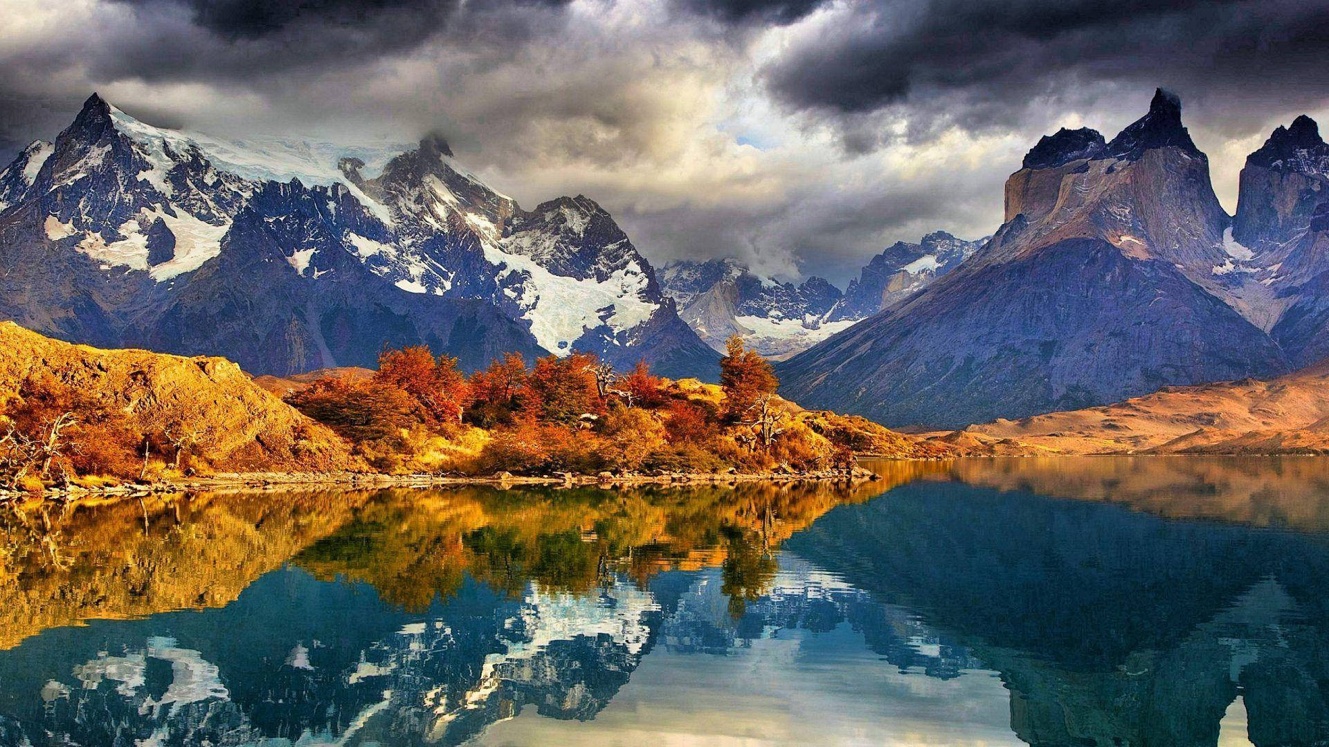Patagonia 桌布 壁紙 — 白雪覆蓋的山脈倒映在平靜的湖面上，周圍是秋葉，天空中有戲劇性的雲彩。