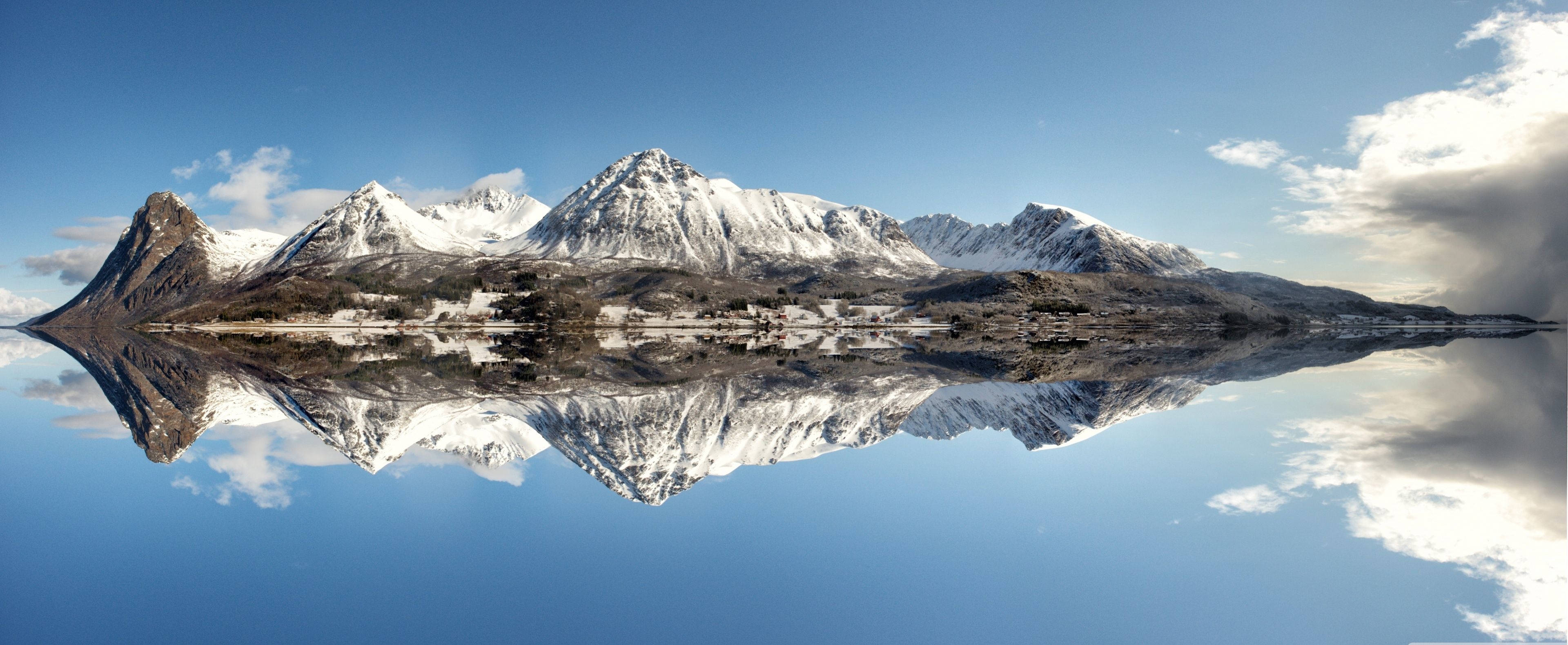 3840x1600 桌布 壁紙 — 白雪覆蓋的山脈和一個小村莊完美地倒映在清澈如鏡的湖面上，天空湛藍。
