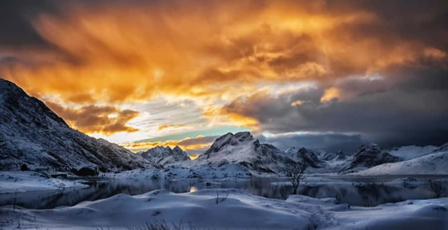 山脈 桌布 壁紙 — 白雪覆蓋的山脈在平靜的湖面上倒映，天空呈現戲劇性的橙色和灰色日落。