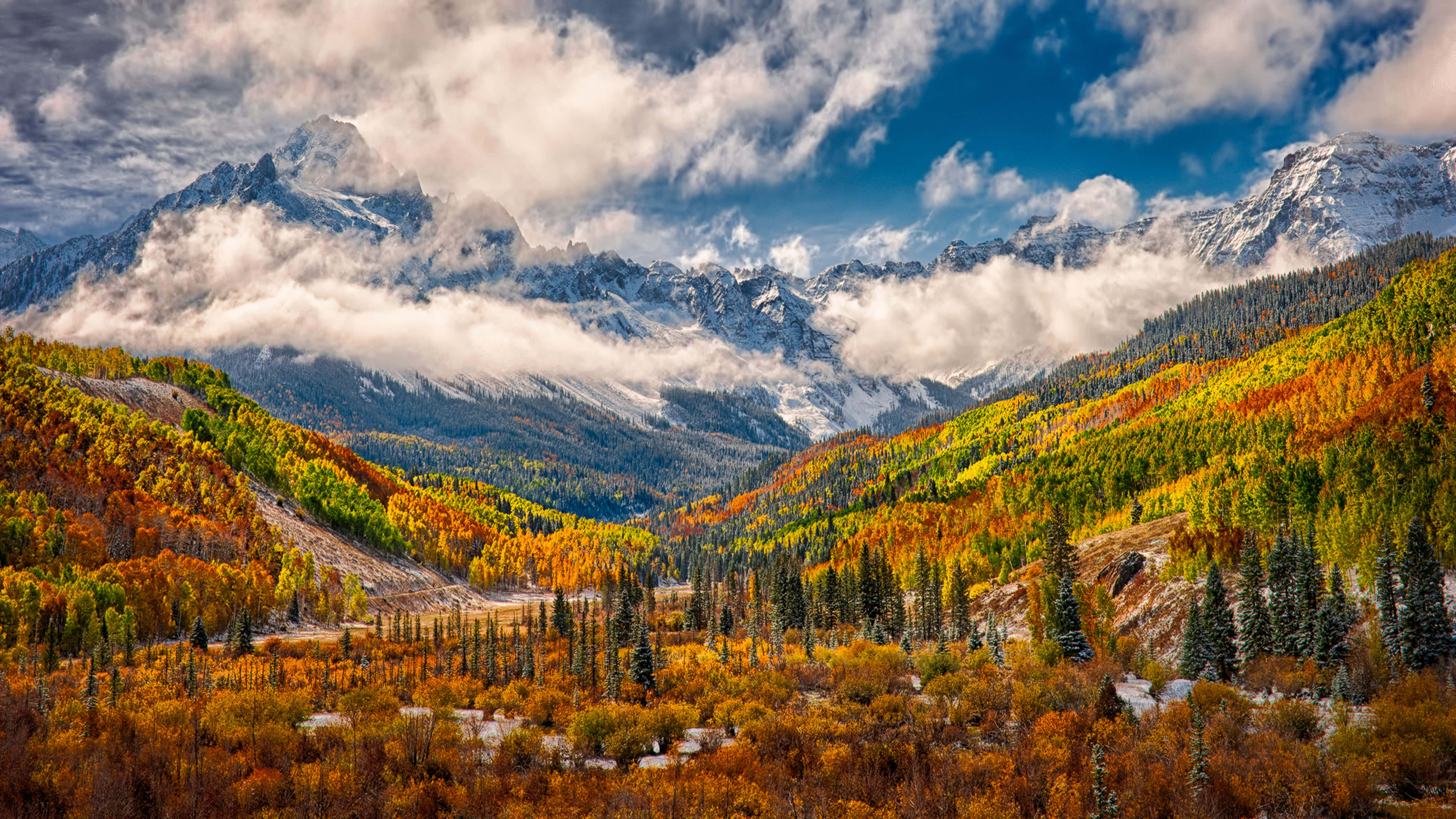 Aspen Colorado 桌布 壁紙 — 白雪覆蓋的山脈在戲劇性的多雲天空下，聳立於充滿鮮豔秋樹的山谷之上。