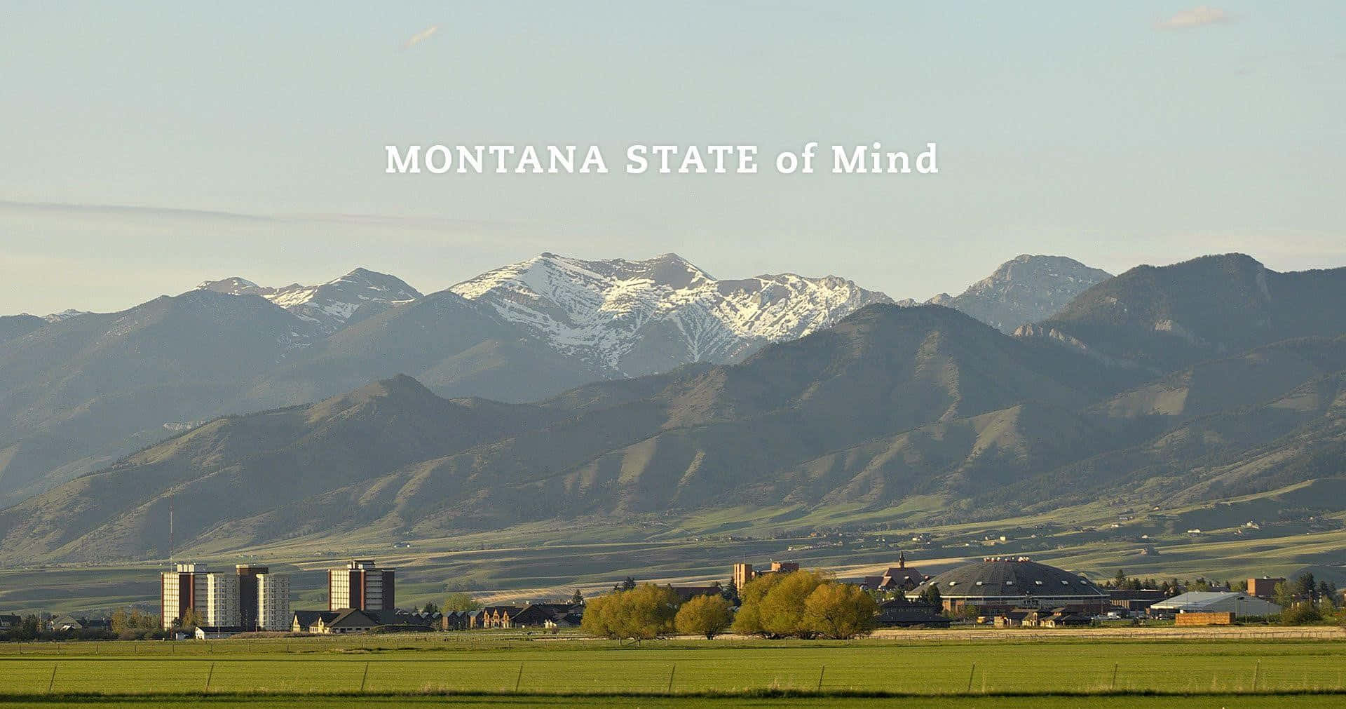 Montana 桌布 壁紙 — 白雪覆蓋的山脈在晴朗天空下，聳立於坐落於廣闊綠色山谷中的小鎮後方。