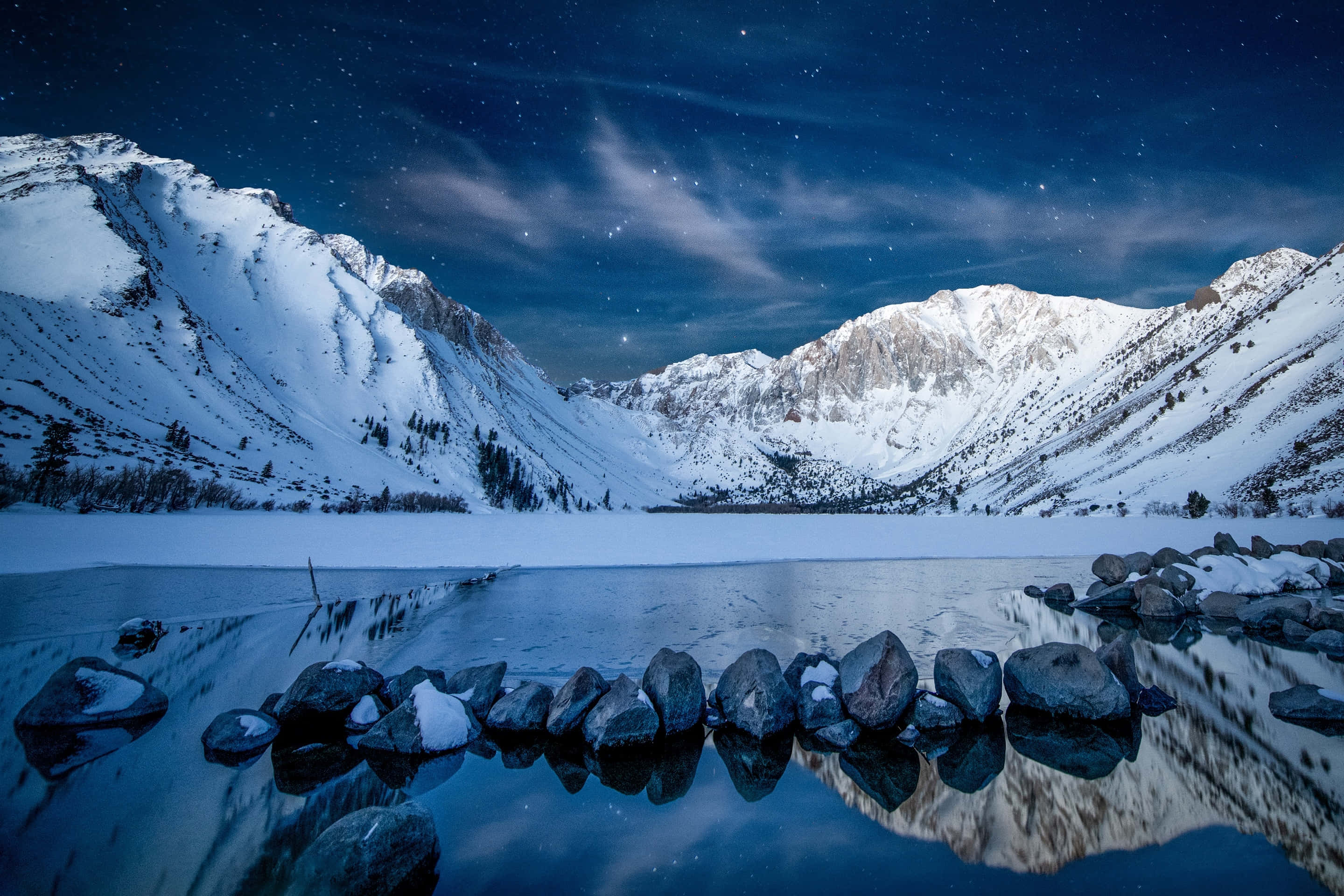 Snowy Mountain 桌布 壁紙 — 白雪覆蓋的山脈在繁星點點的夜空下完美地倒映在冰凍的湖面上，周圍環繞著深色的岩石。