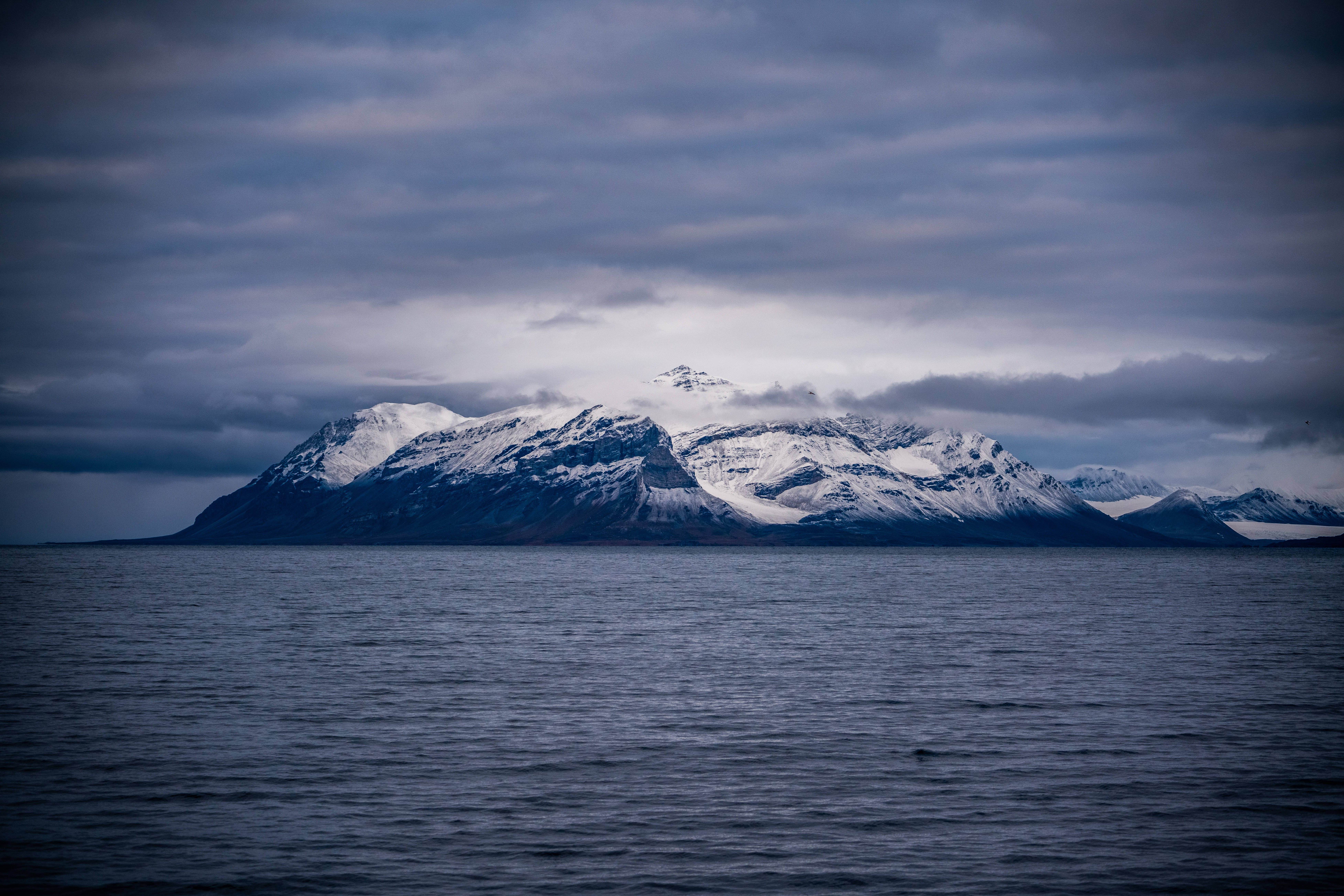 Svalbard 桌布 壁紙 — 白雪覆蓋的山脈在陰沉多雲的天空下從平靜的水面上戲劇性地升起。