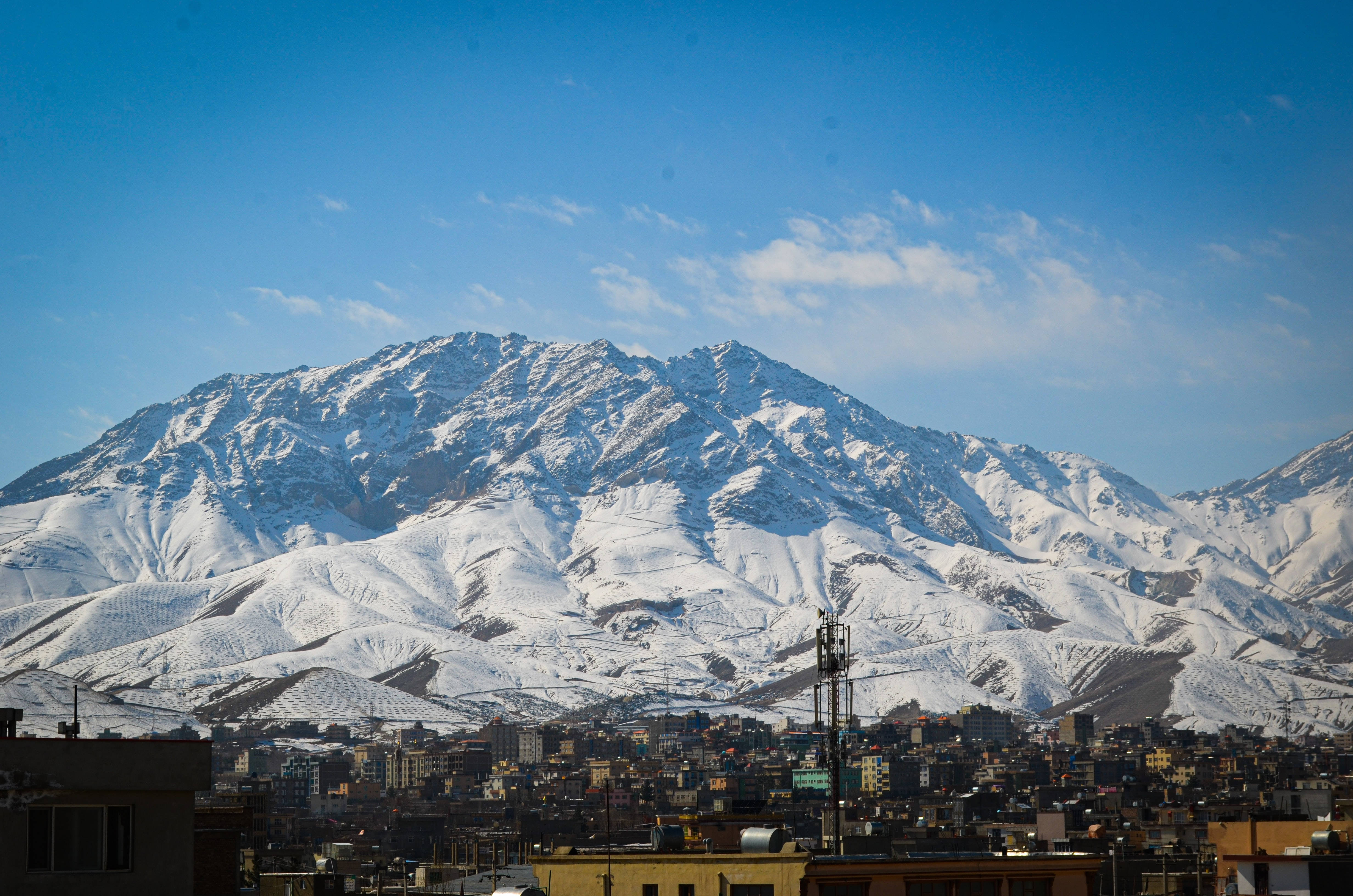 Kabul 桌布 壁紙 — 白雪覆蓋的山脈聳立於一座廣闊城市之上，天空湛藍清澈，飄著縷縷薄雲。