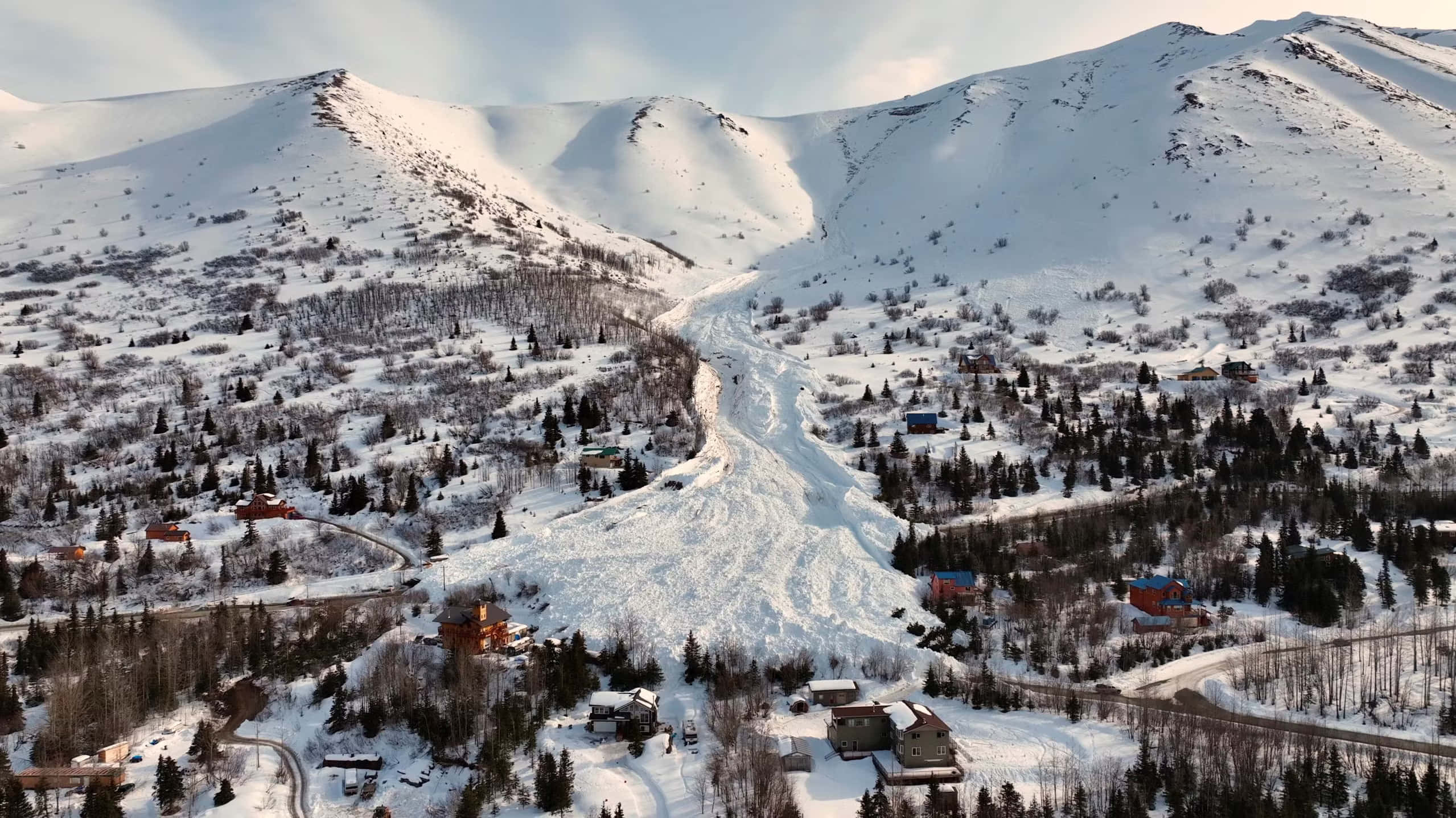 Snowy Mountain 桌布 壁紙 — 白雪覆蓋的山谷鳥瞰圖，蜿蜒的滑雪道和散落在常綠樹林中的小屋。