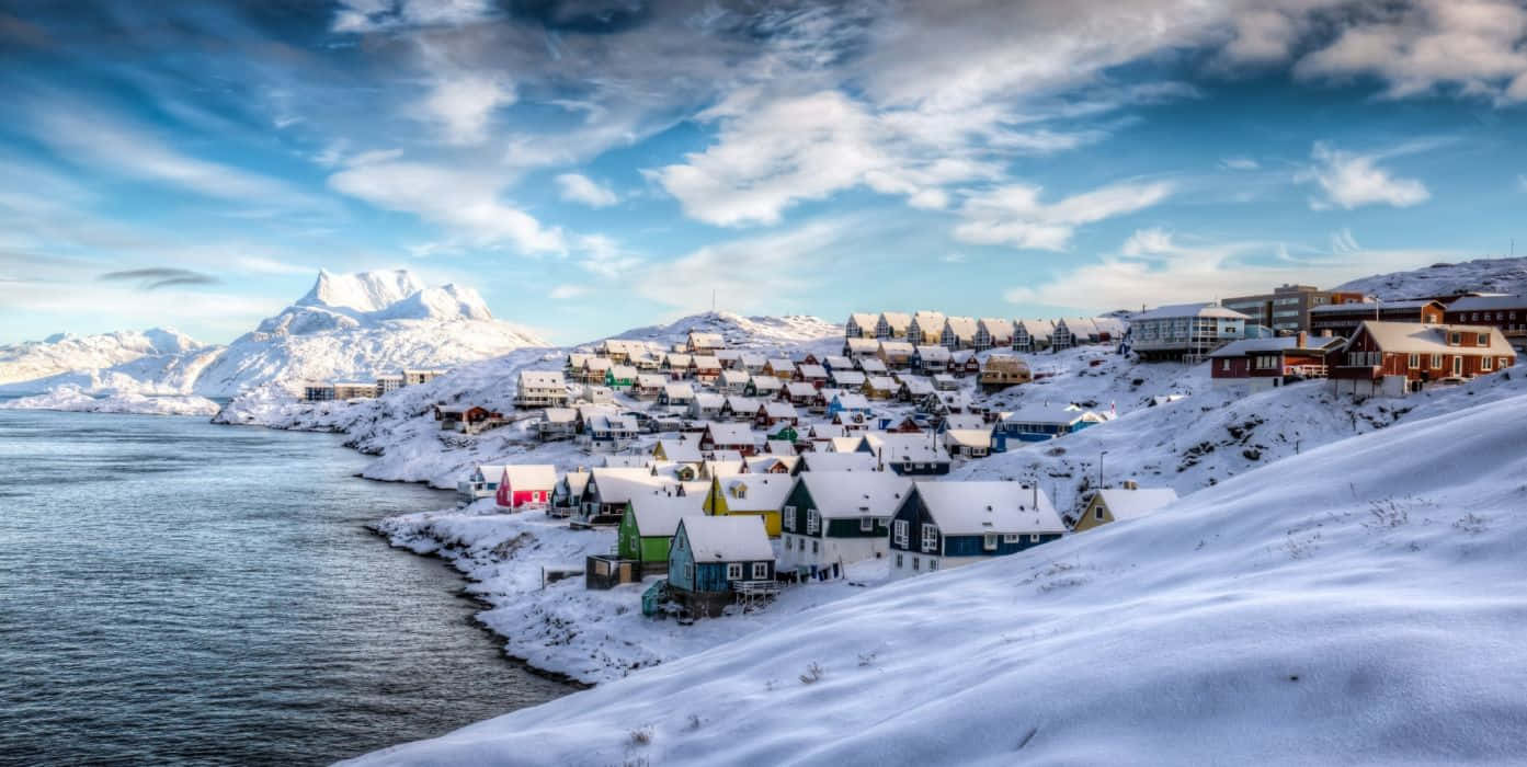 Greenland 桌布 壁紙 — 白雪覆蓋的彩色房屋依附在積雪的山坡上，旁邊是平靜的峽灣，上方是帶有縷縷白雲的明亮藍天。