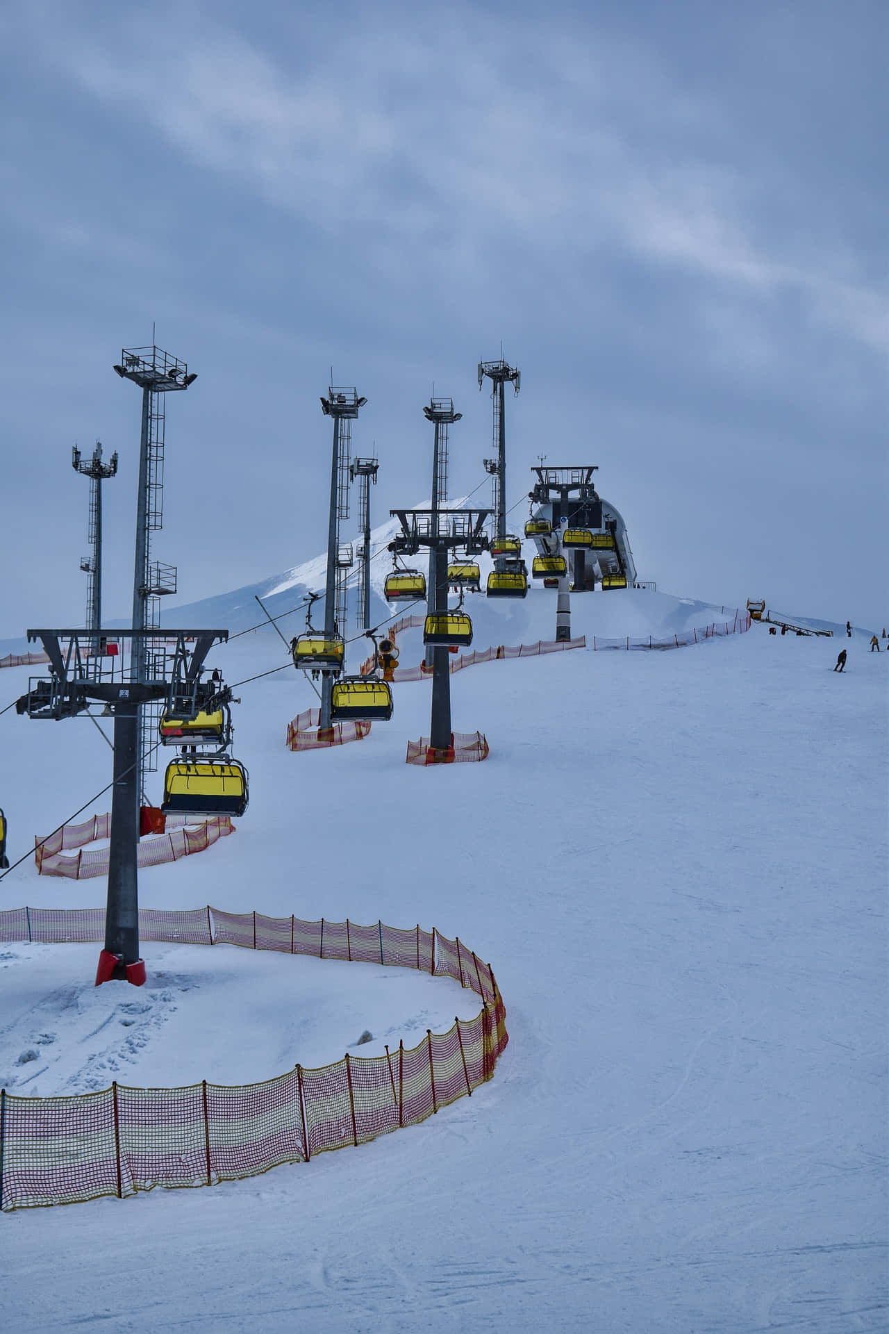 Ski Resort 桌布 壁紙 — 白雪覆蓋的滑雪坡道上，黃色纜車在陰天上升，遠處的山丘上散落著滑雪者。